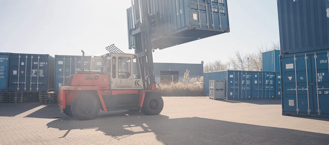 Forklift lifting a blue shipping container in a yard with stacked containers around.