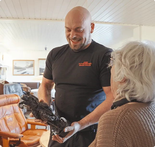 Smiling man shows a dark horse statue to an elderly person with white hair indoors.