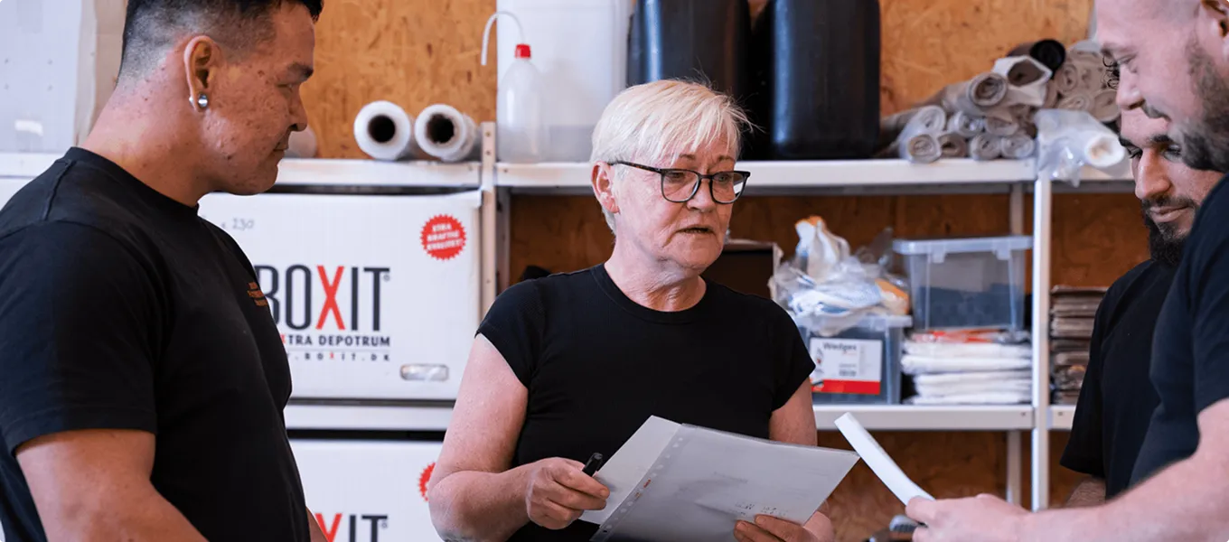 Older woman discussing documents with three men in a workshop with shelves of supplies behind them.
