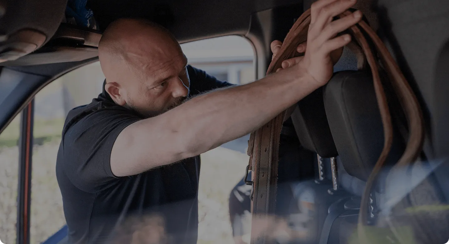 Man in a black shirt securing a brown belt or strap inside a vehicle near the headrest.