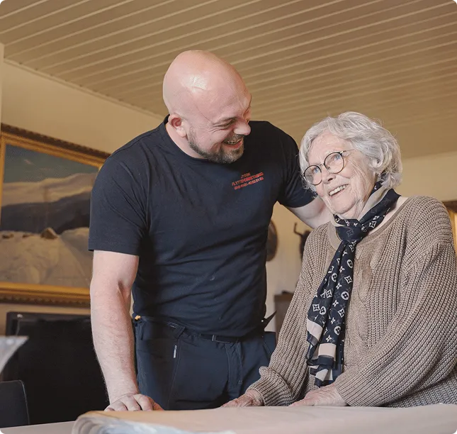 Smiling man with arm around elderly woman wearing glasses and scarf at a table indoors.