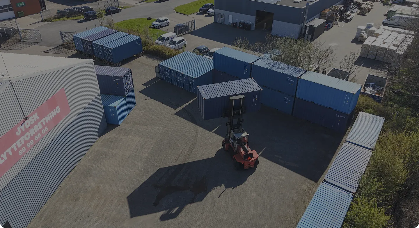 Forklift lifting a blue shipping container in an industrial yard with stacked containers and parked cars.