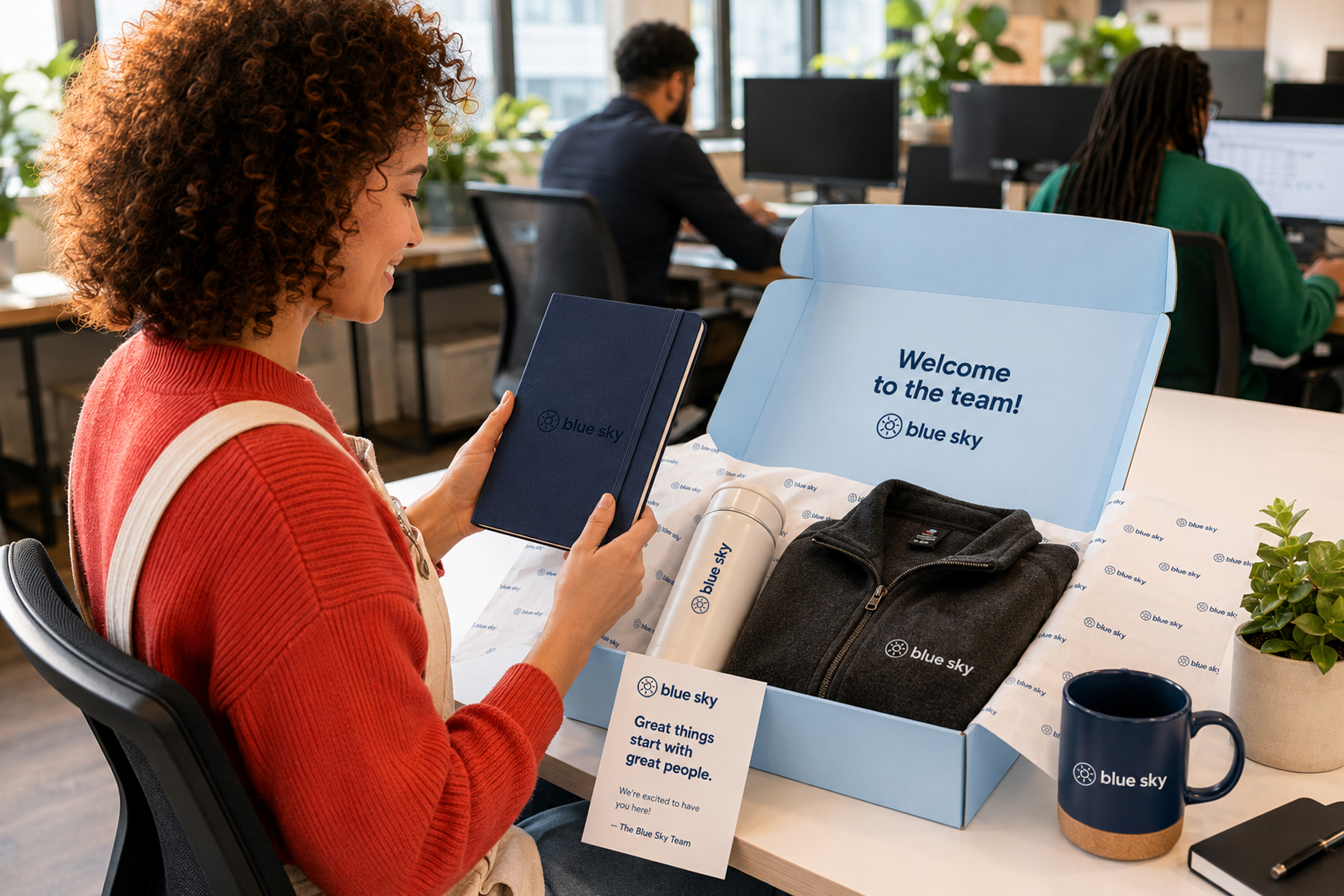 New hire reading a Blue Sky notebook beside an open welcome box with printed tissue, a welcome card, water bottle, jacket, and mug