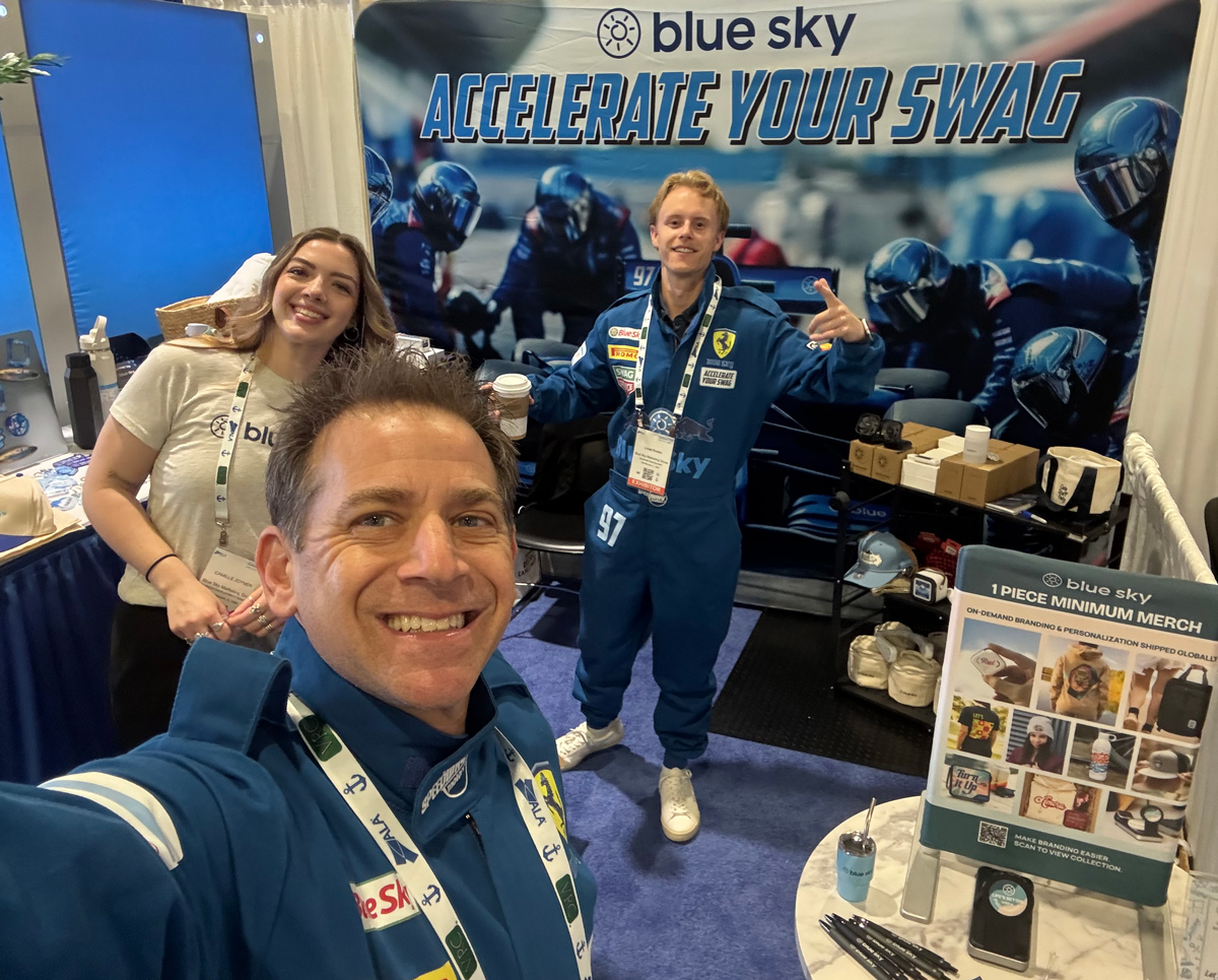 Blue Sky team members, two in racing suits, taking a selfie at the Blue Sky Accelerate Your Swag trade show booth