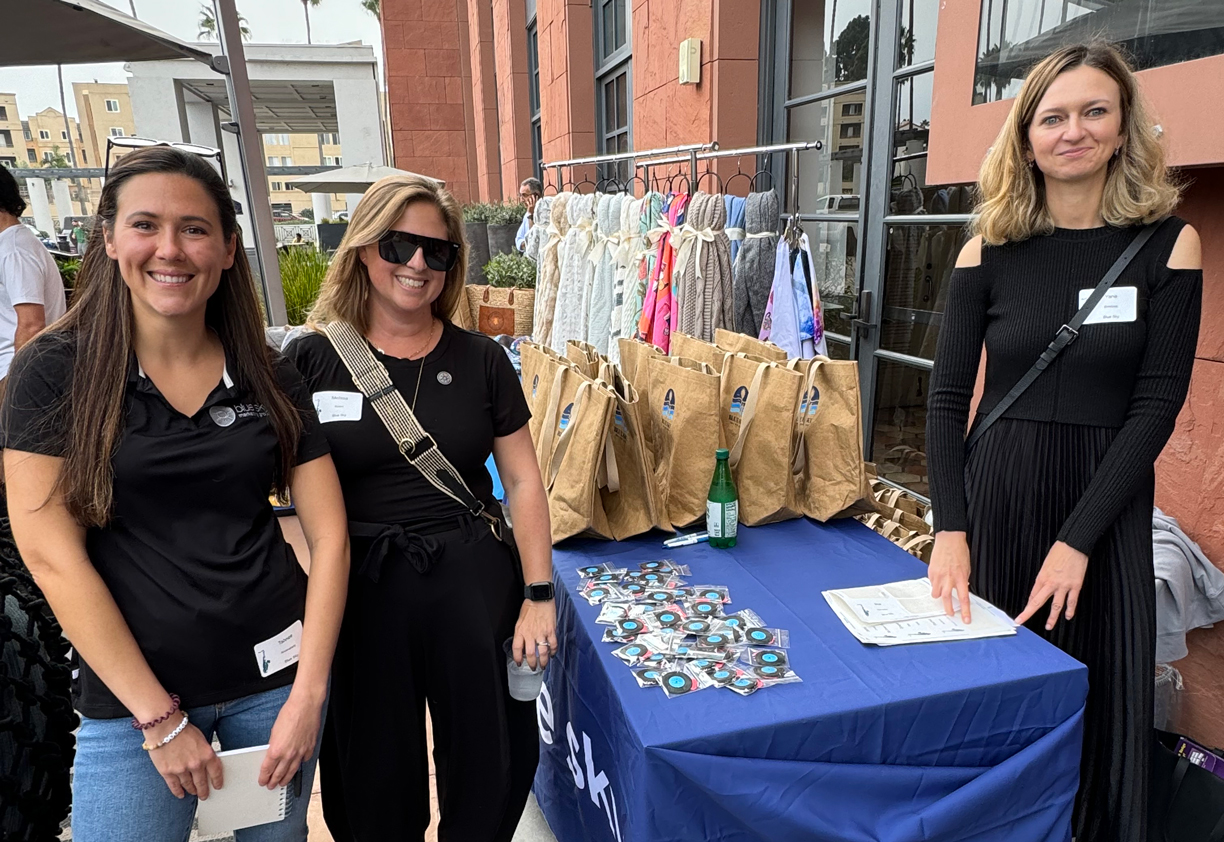 Three Blue Sky team members smiling beside an outdoor event table set with branded tote bags and giveaways