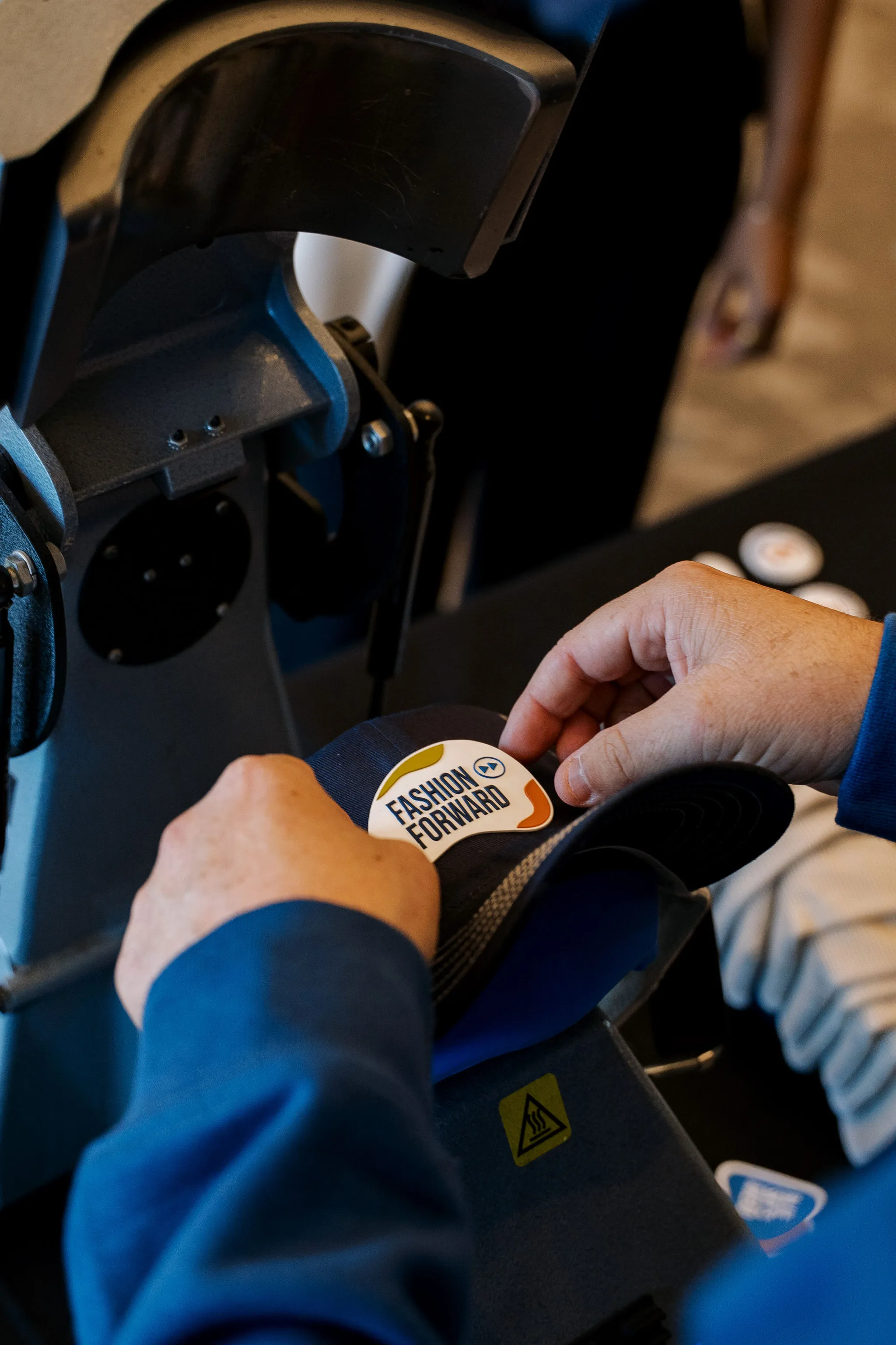 Hands pressing a Fashion Forward patch onto a cap at a live heat-press customization station