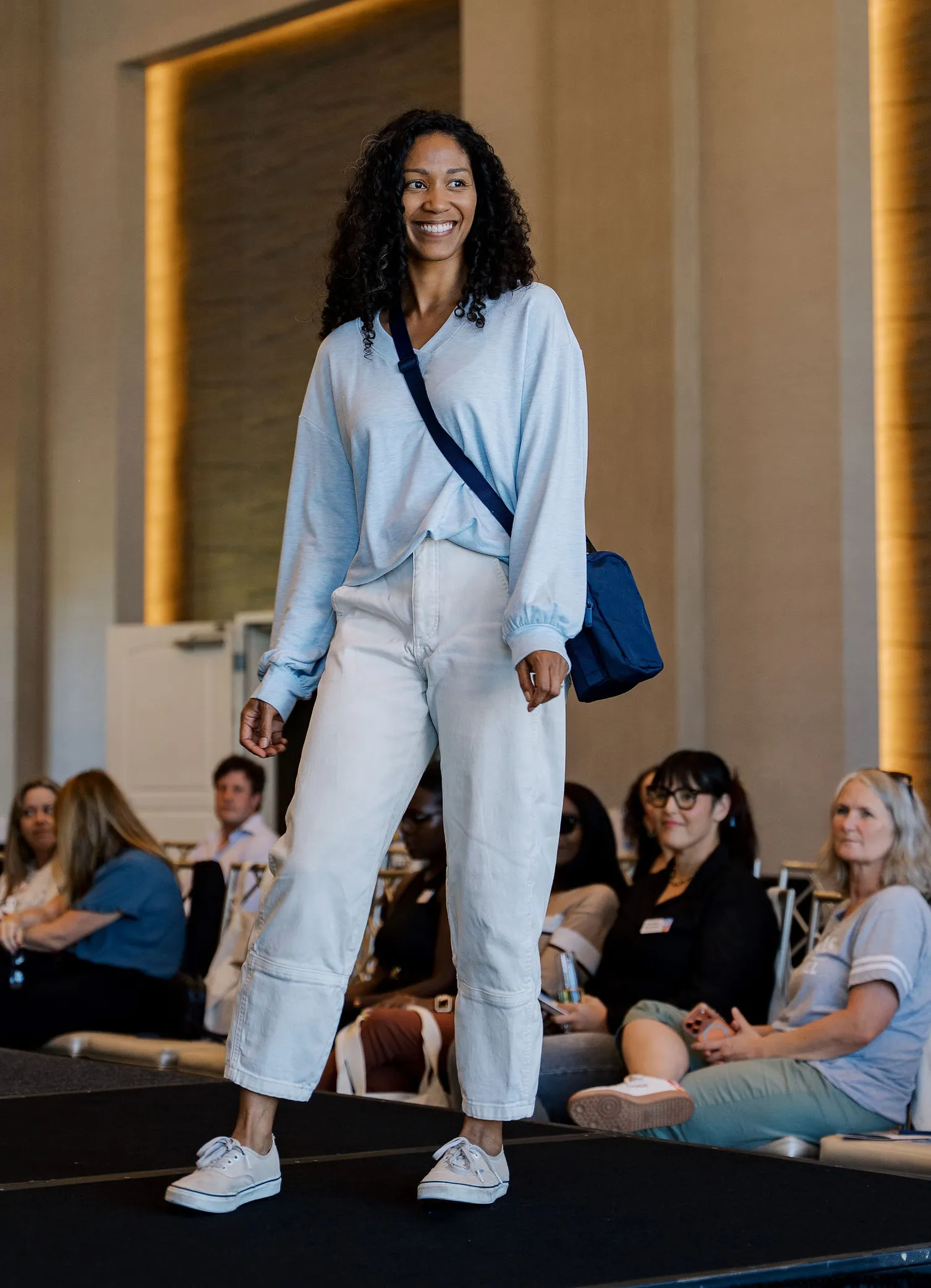Model smiling on the Fashion Forward runway in a light blue top, white pants, and a navy crossbody bag as guests watch
