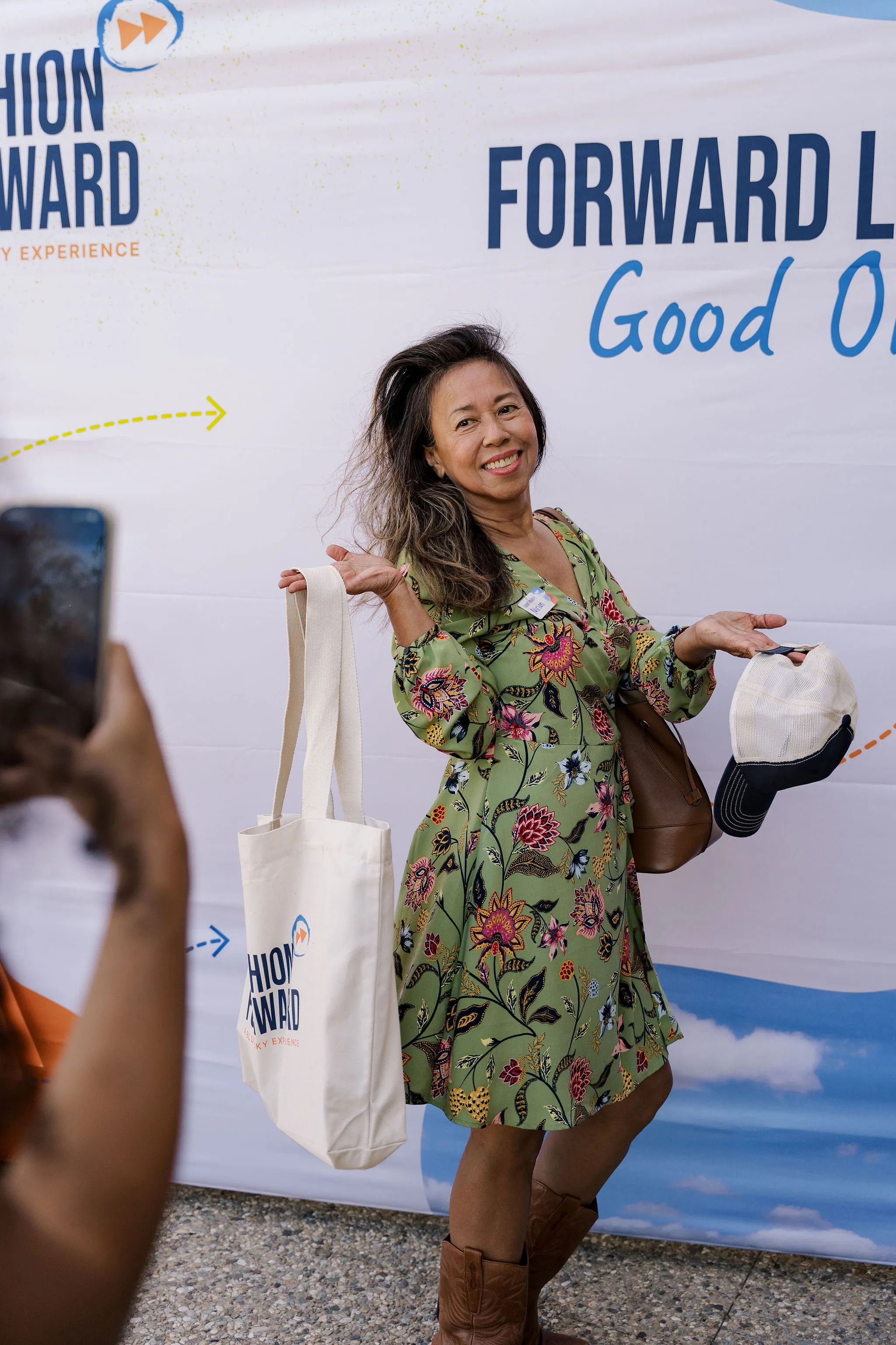 Guest in a green floral dress posing with a Fashion Forward tote and cap at the Forward Looks photo backdrop