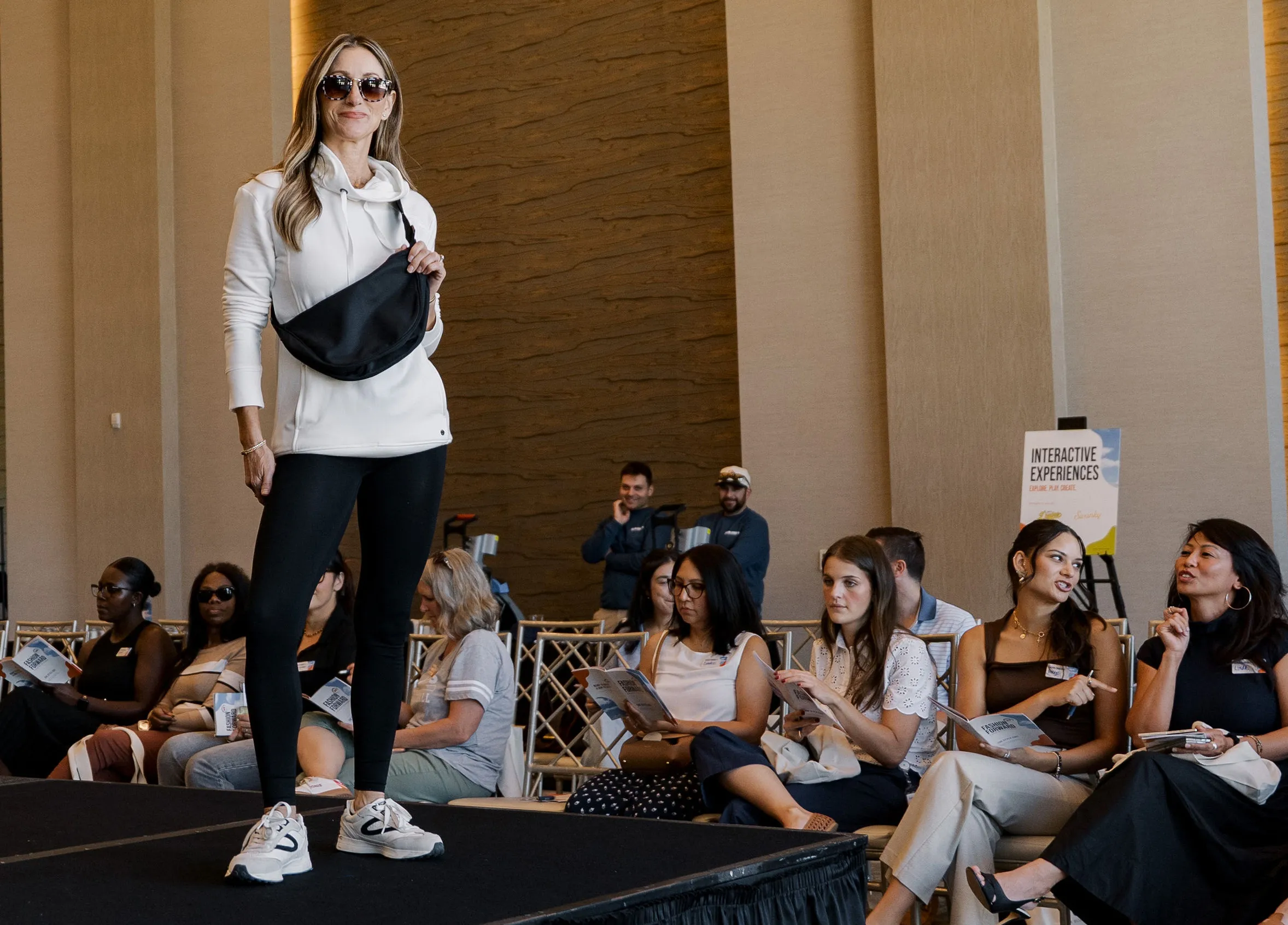 Model in a white zip hoodie and black crossbody bag walking the Fashion Forward runway as guests look on