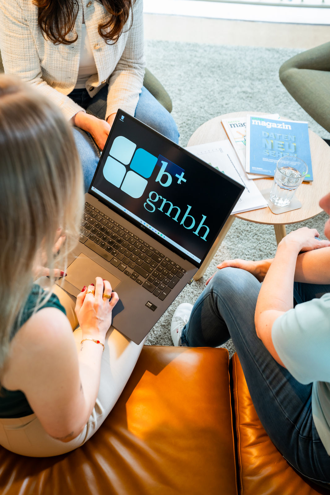 Person arbeitet am Laptop mit b+ GmbH Logo, im Hintergrund eine Büro-Lounge.
