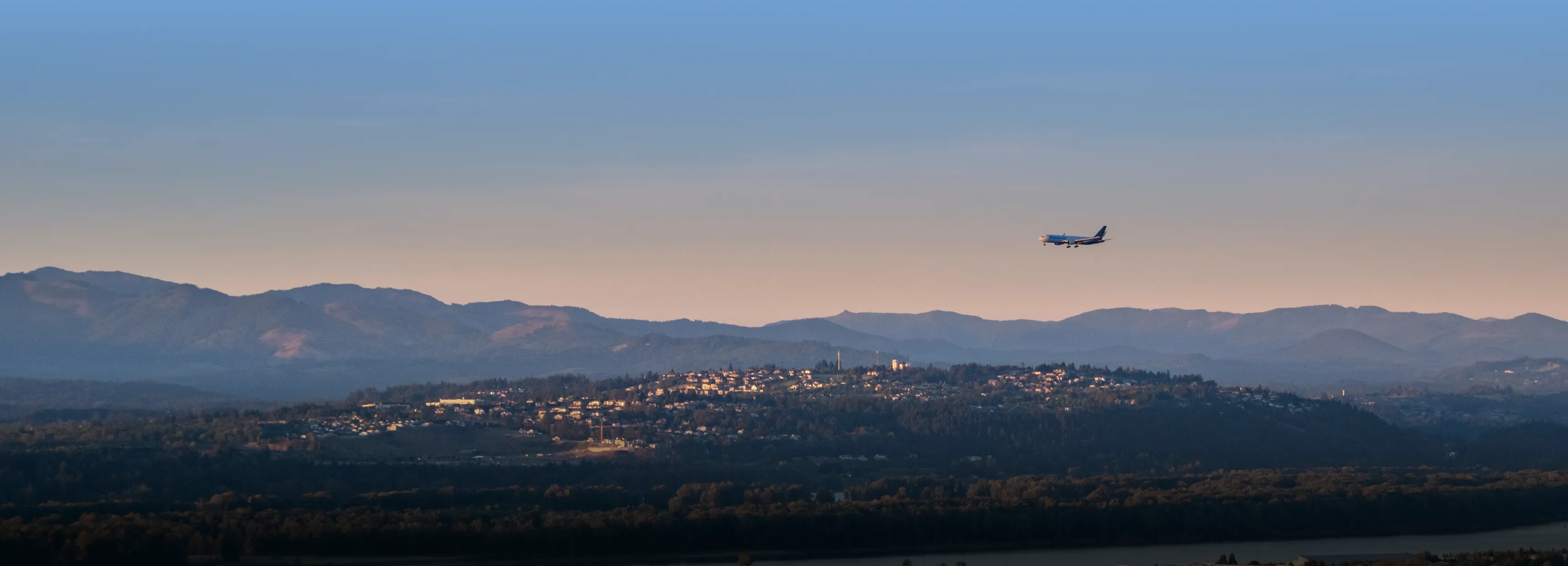 Airplane flying over hills with houses under a clear sky at dusk.