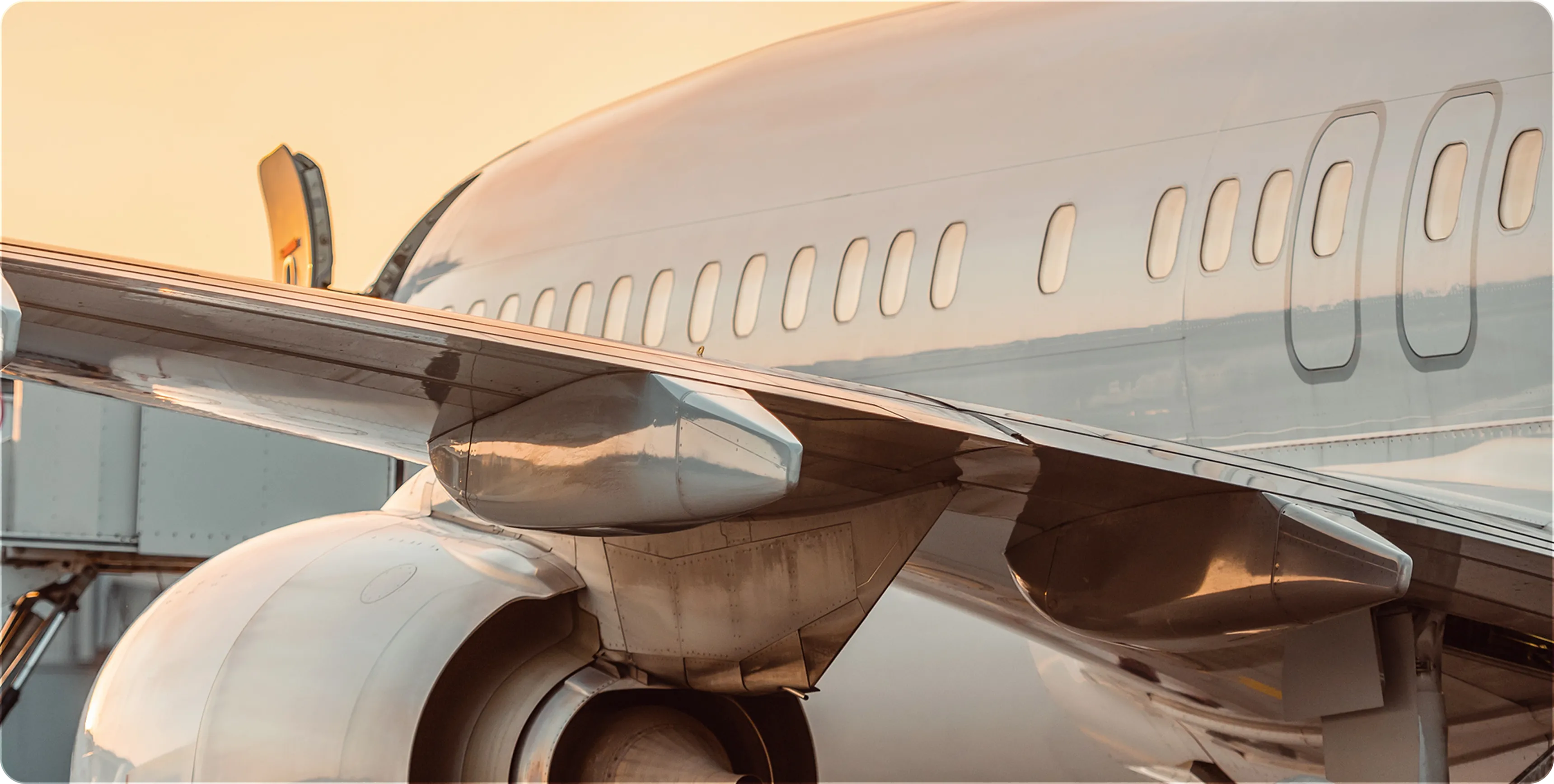 Close-up of airplane wing and jet engine with passenger windows and boarding bridge in background.