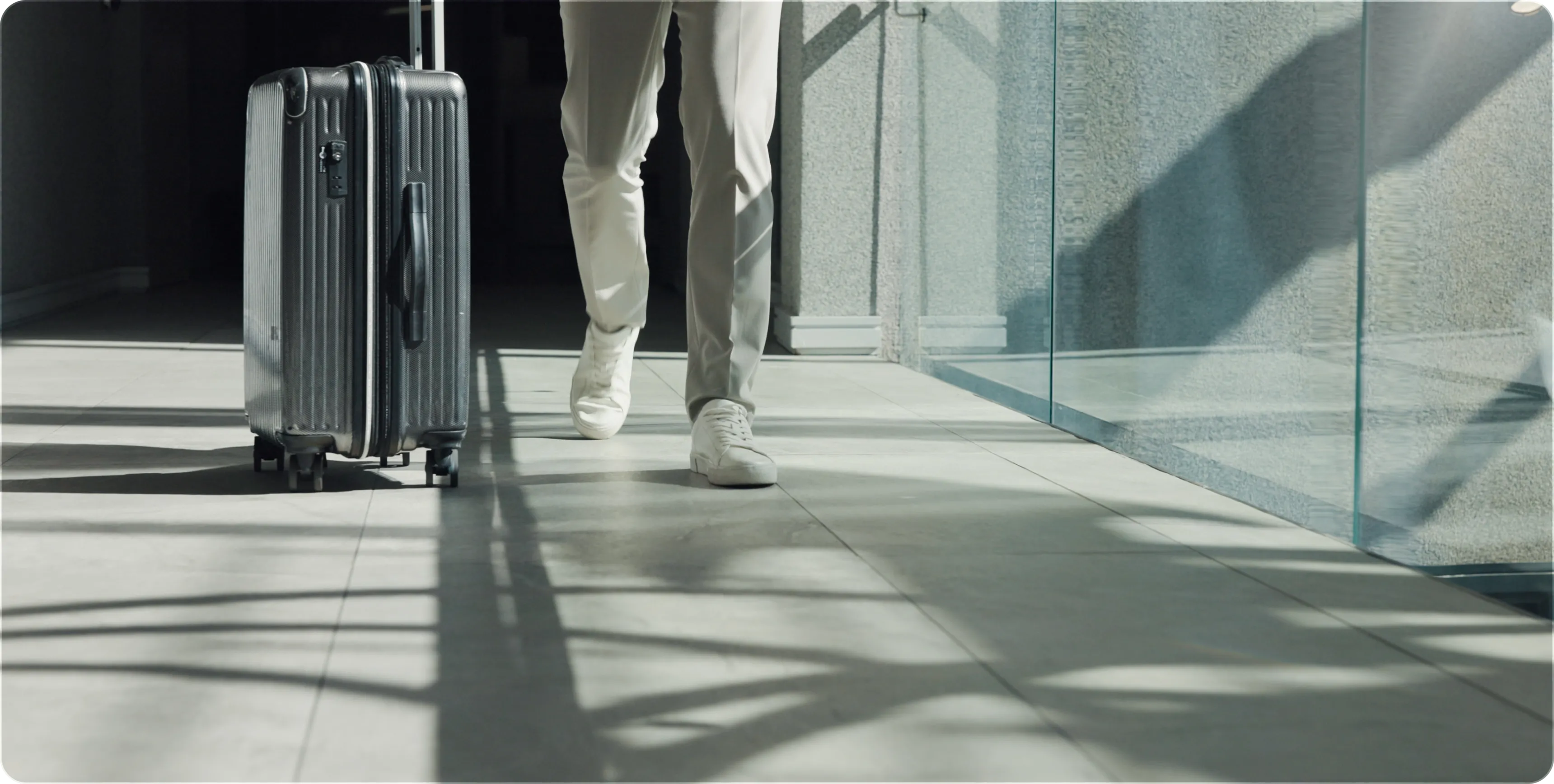 Person walking next to a black rolling suitcase on a tiled floor with glass walls nearby.