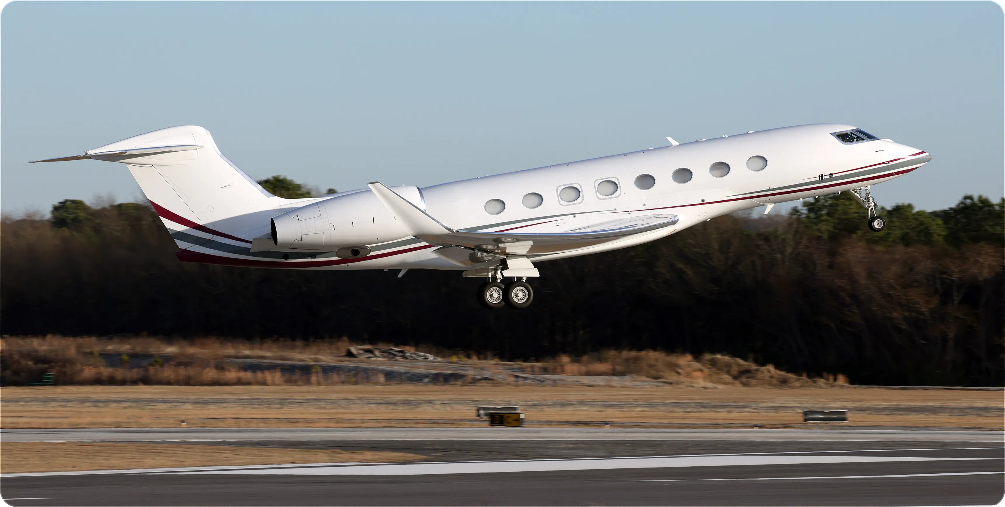 White private jet with red and gray stripes taking off from runway near trees.