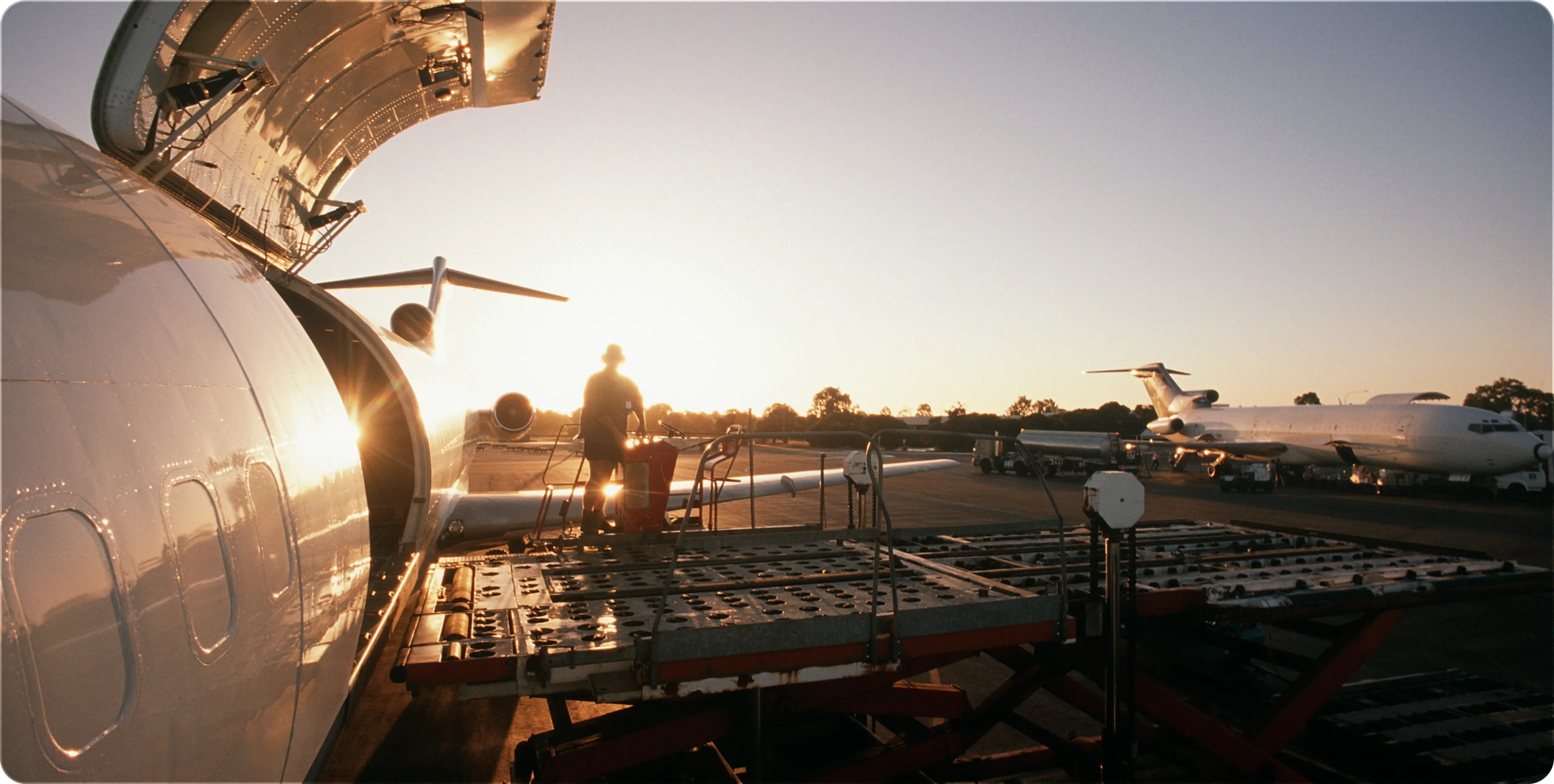 Person working on cargo loading platform next to an airplane at sunset on an airport tarmac.
