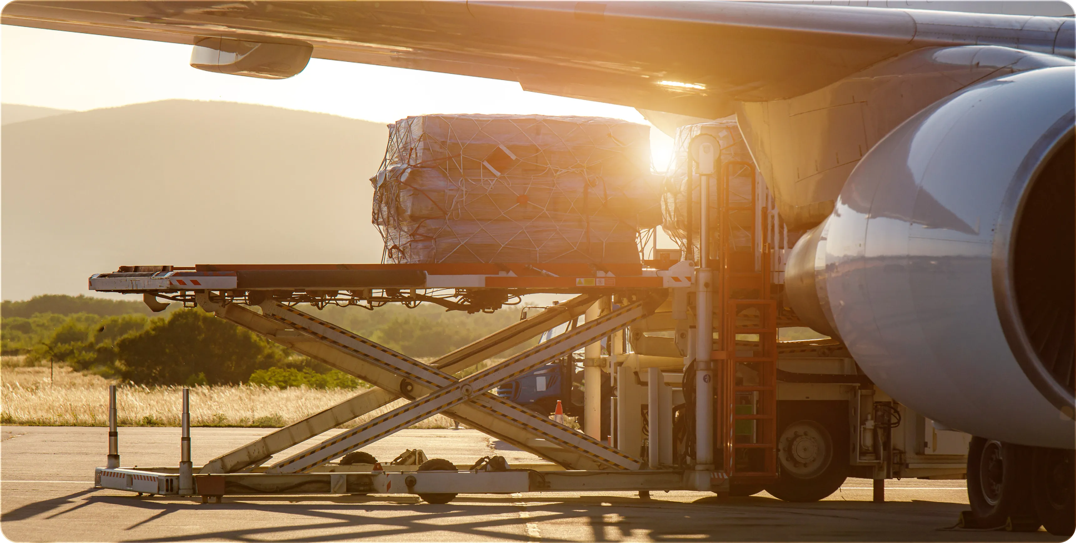 Cargo being loaded into an airplane using a scissor lift platform at sunset on a tarmac.