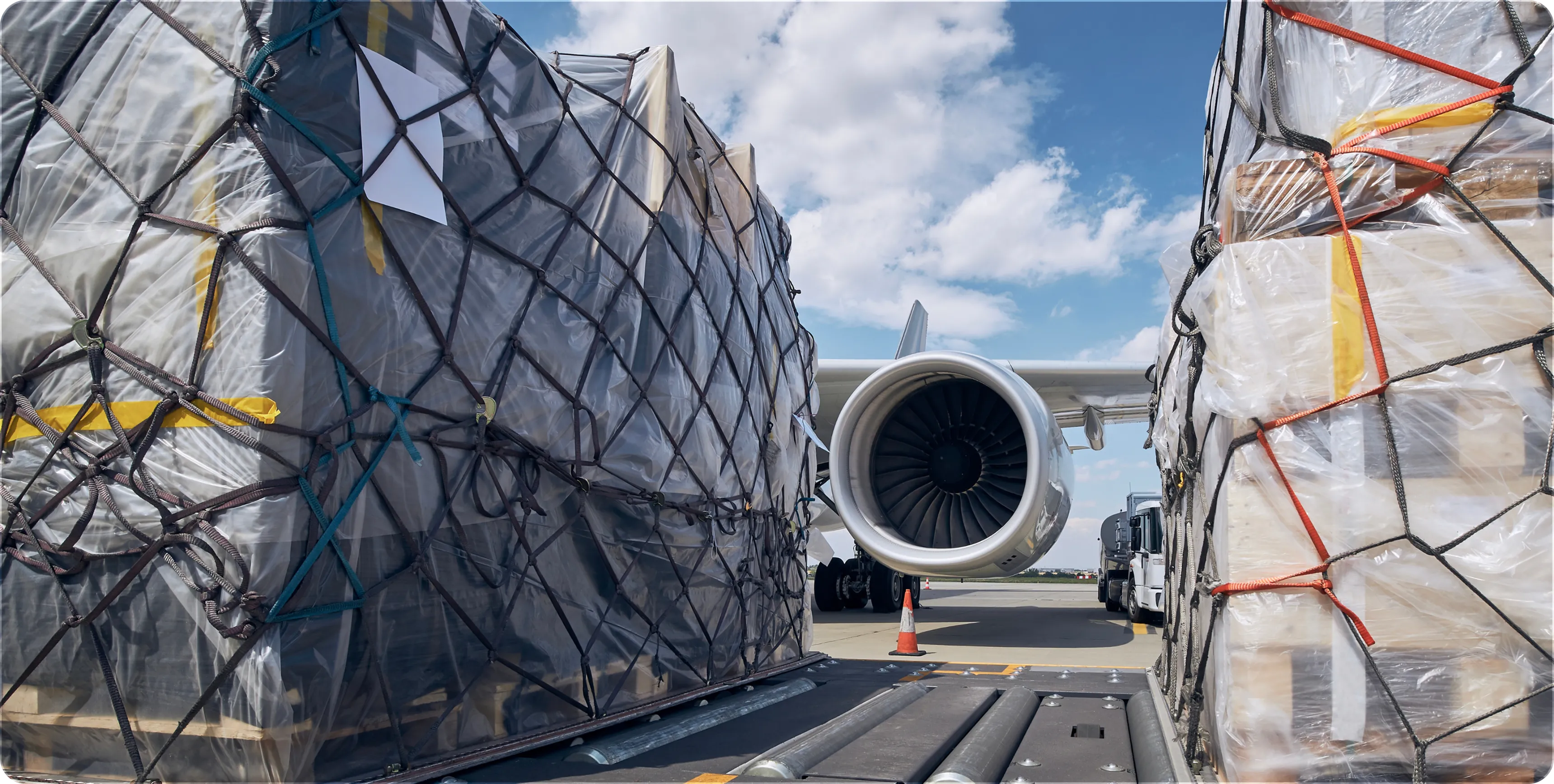 Cargo wrapped in plastic and netting on a conveyor with an airplane engine and truck in background at airport.