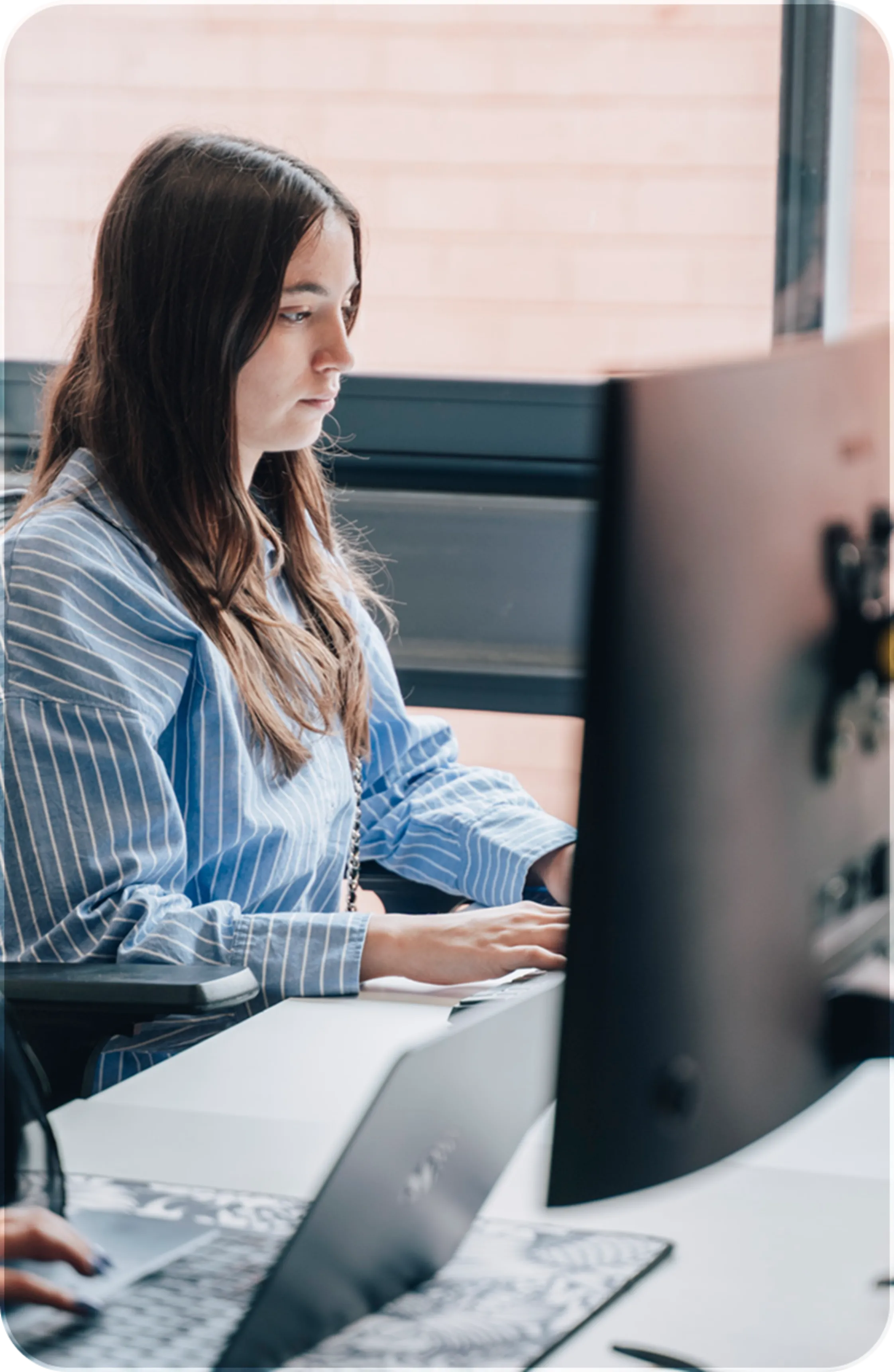 Woman in blue striped shirt working on a desktop computer at an office desk.