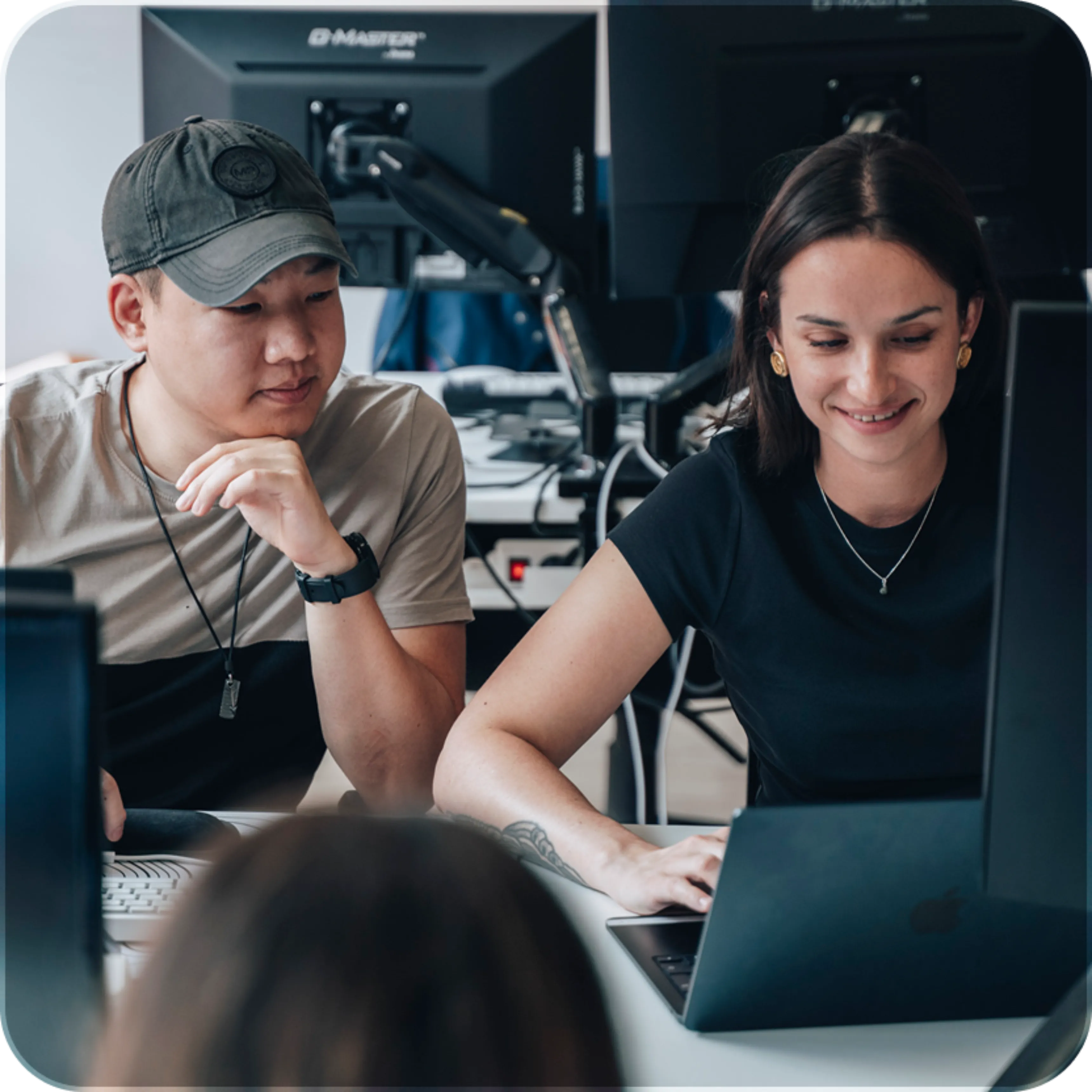 A man and a woman working together on a laptop in an office with computer monitors in the background.