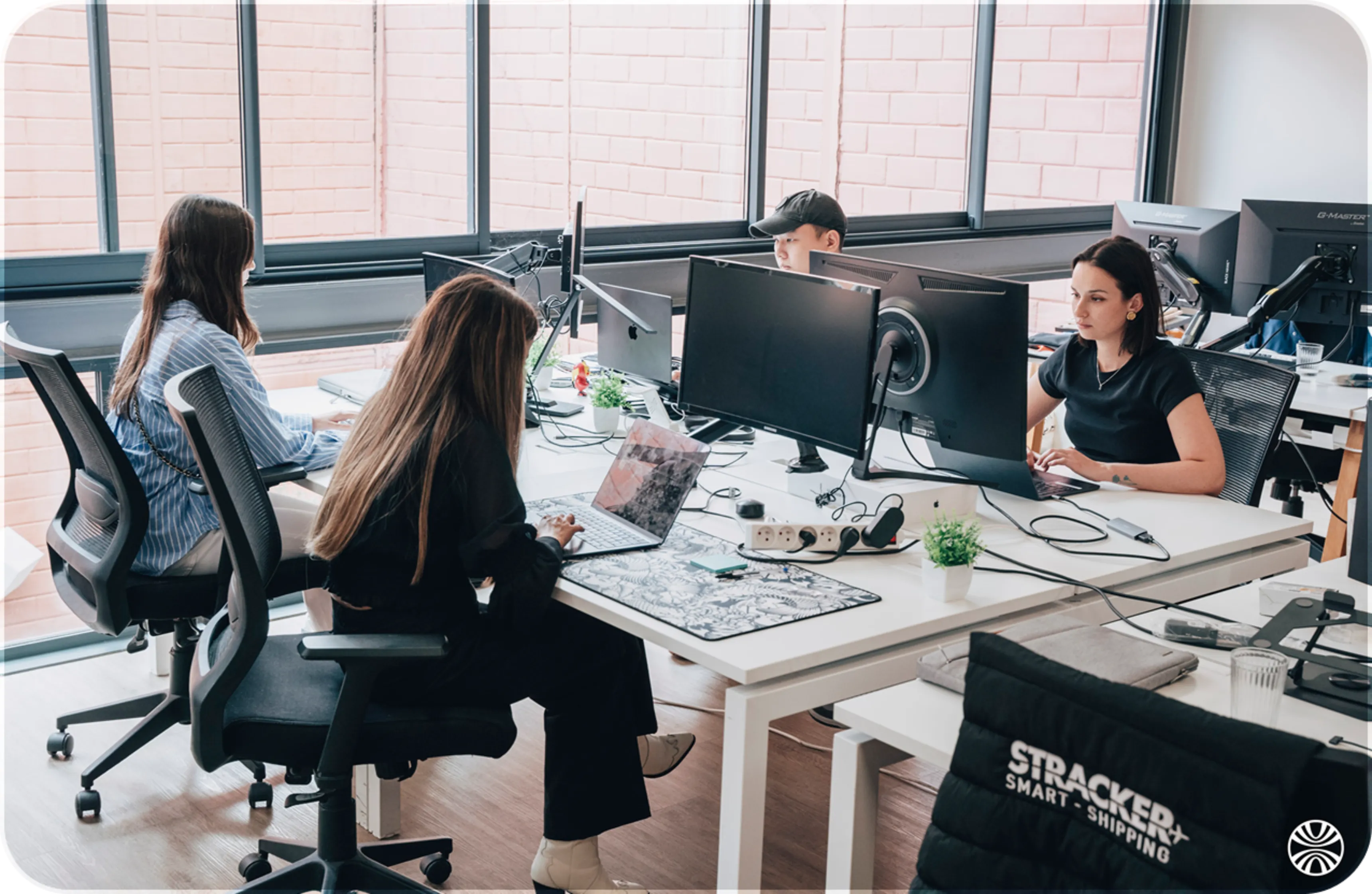 Four people working on computers at a shared office desk with large windows in the background.