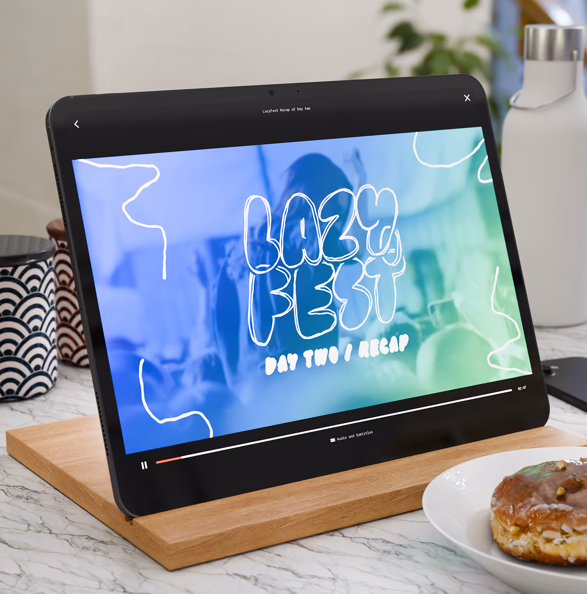 A LazyType Mockup with an iPad on a wooden stand showing a video titled 'Lazy Fest Day Two Recap' on a kitchen counter.