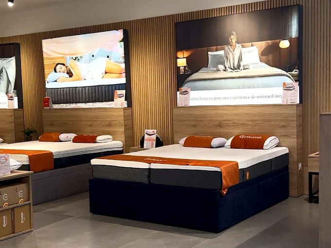Modern bedroom showroom with Emma mattress, orange pillows, and a large framed photo on wooden floor.