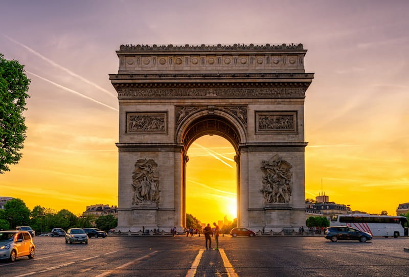 Arc de Triomphe à Paris au coucher du soleil avec des voitures et des piétons autour.