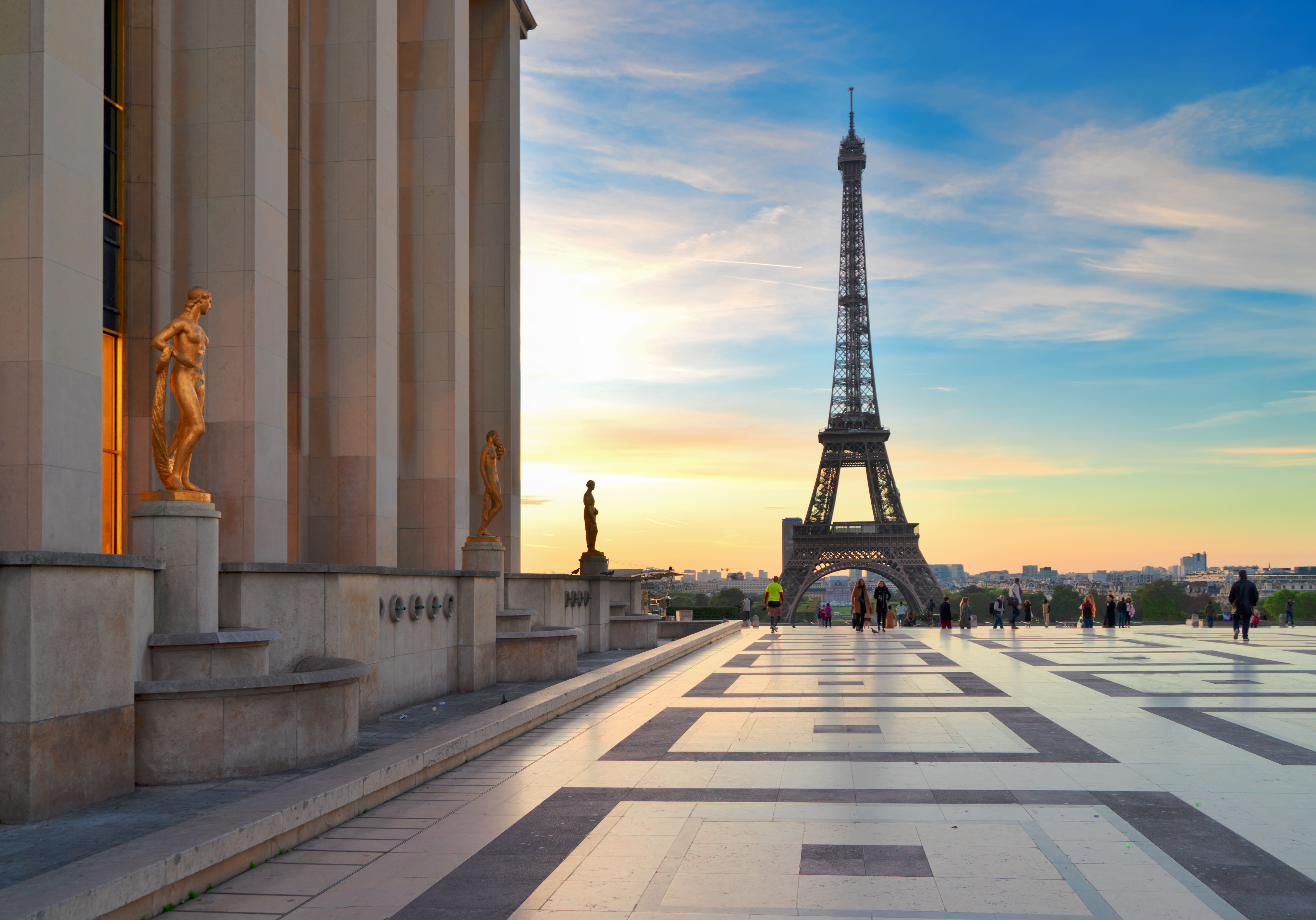 Vue de la Tour Eiffel au coucher du soleil depuis le parvis avec des statues dorées et des personnes se promenant.