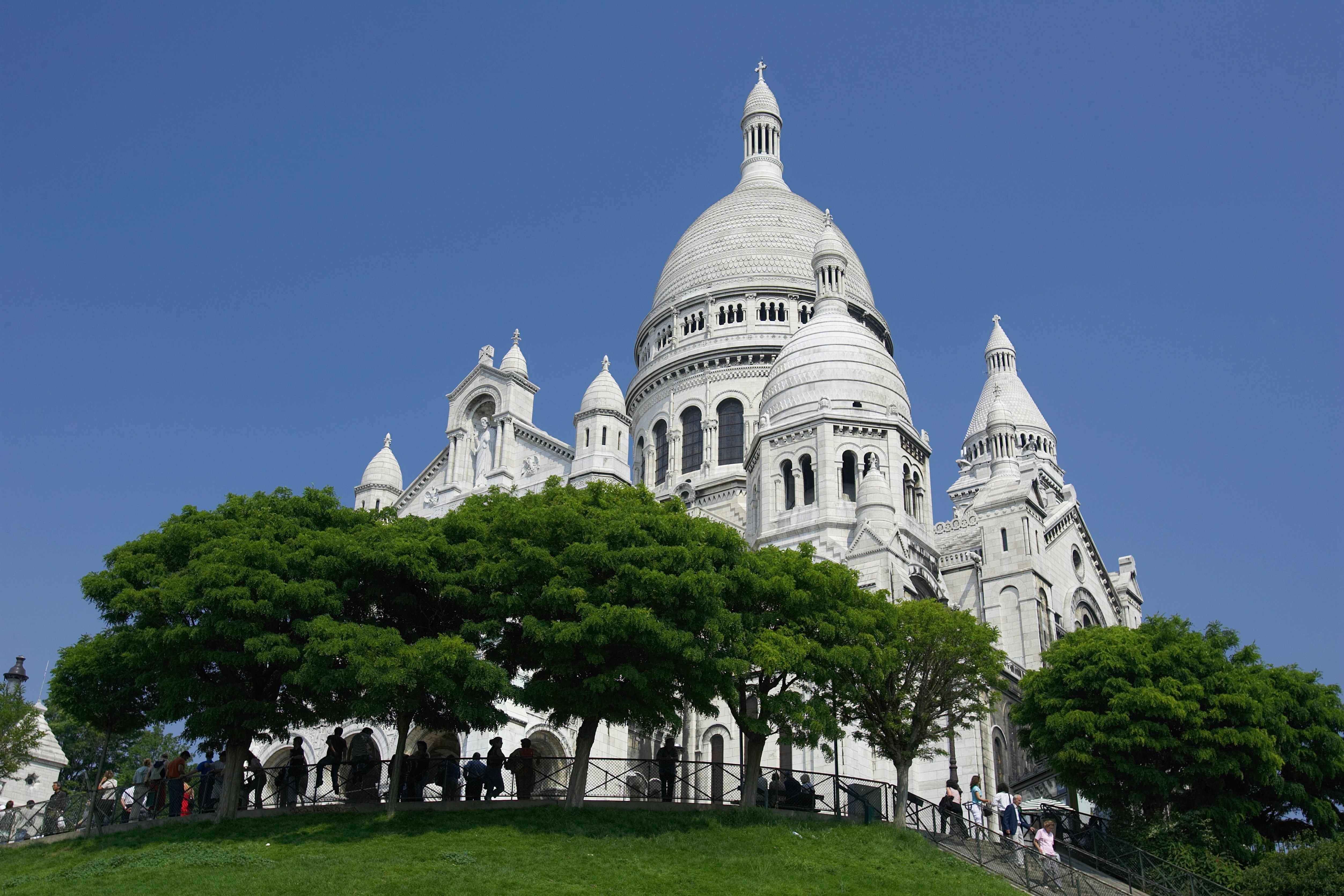 Le Sacré-Cœur de Montmartre, une basilique blanche avec des dômes emblématiques sous un ciel bleu clair, entourée d'arbres verts.