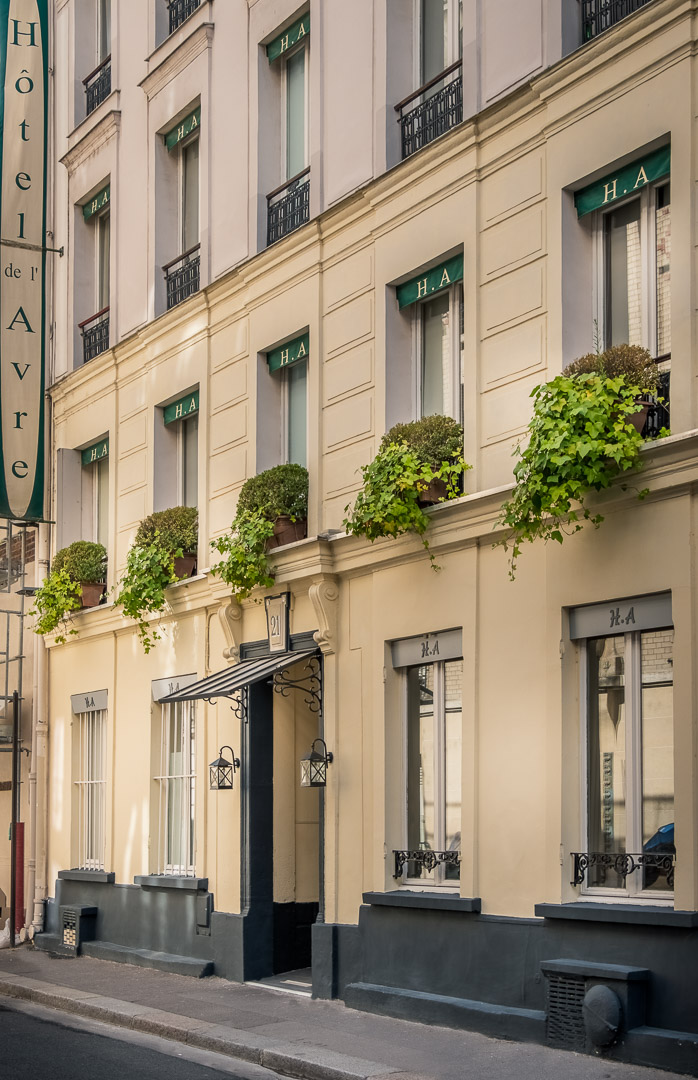 Façade beige élégante de l'hôtel Avre avec de grandes fenêtres ornées de plantes vertes en pots, un auvent noir au-dessus de la porte d'entrée centrale numérotée 21, et une enseigne verticale indiquant Hôtel de l'Avre.