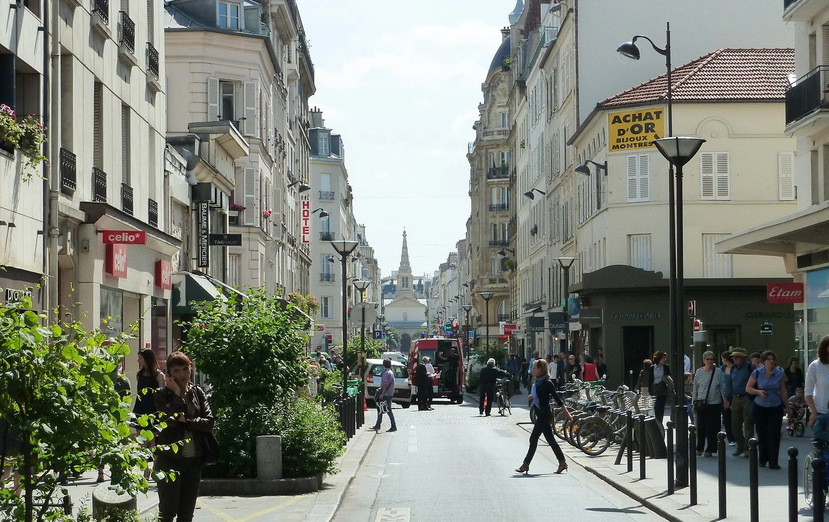 Rue commerçante animée avec des piétons, des vélos en stationnement, des magasins aux façades anciennes, et une église au fond sous un ciel dégagé.