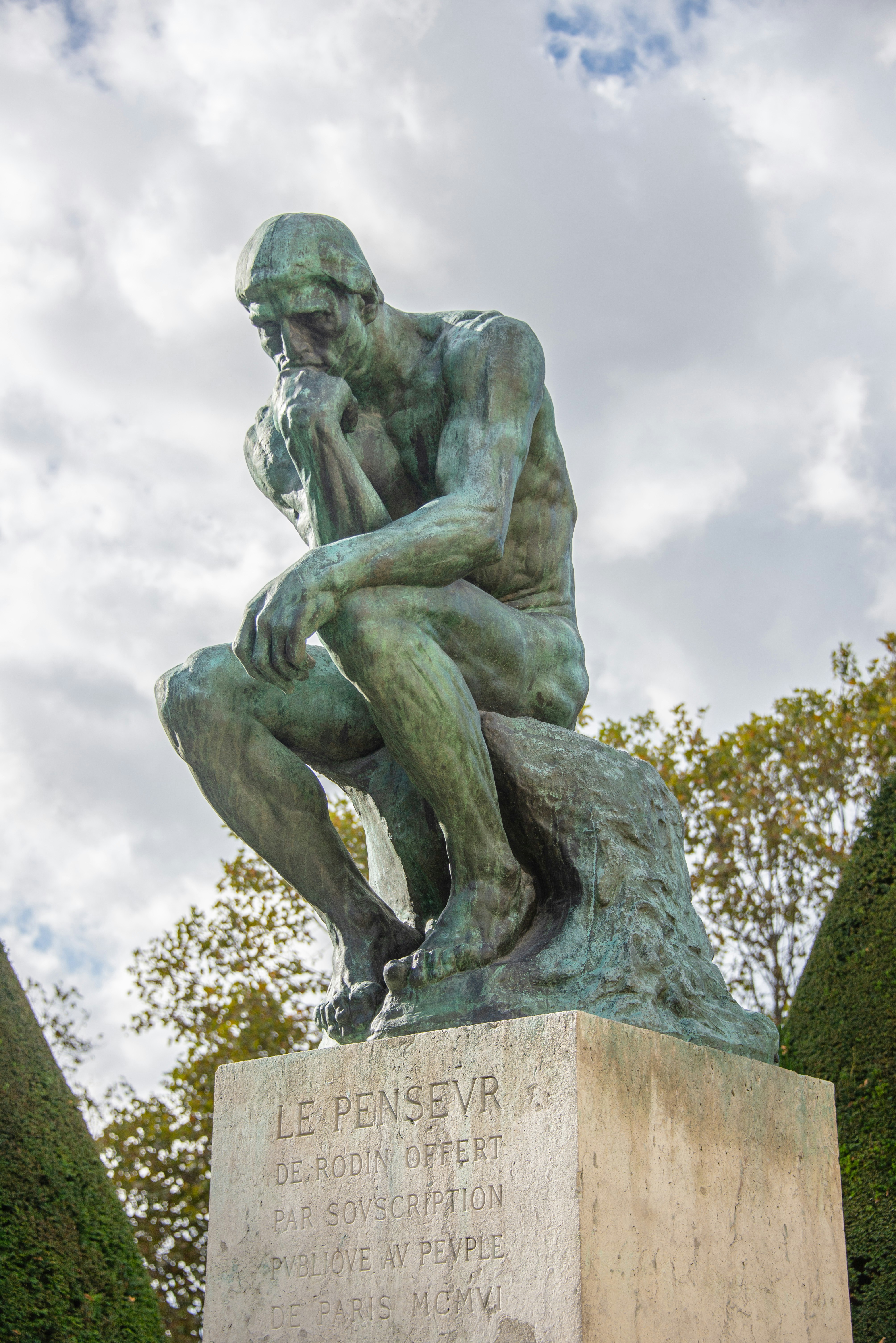 Statue en bronze de Le Penseur d'Auguste Rodin, représentant un homme nu en position assise, le menton reposant sur sa main droite, entouré d'arbres et sous un ciel nuageux.