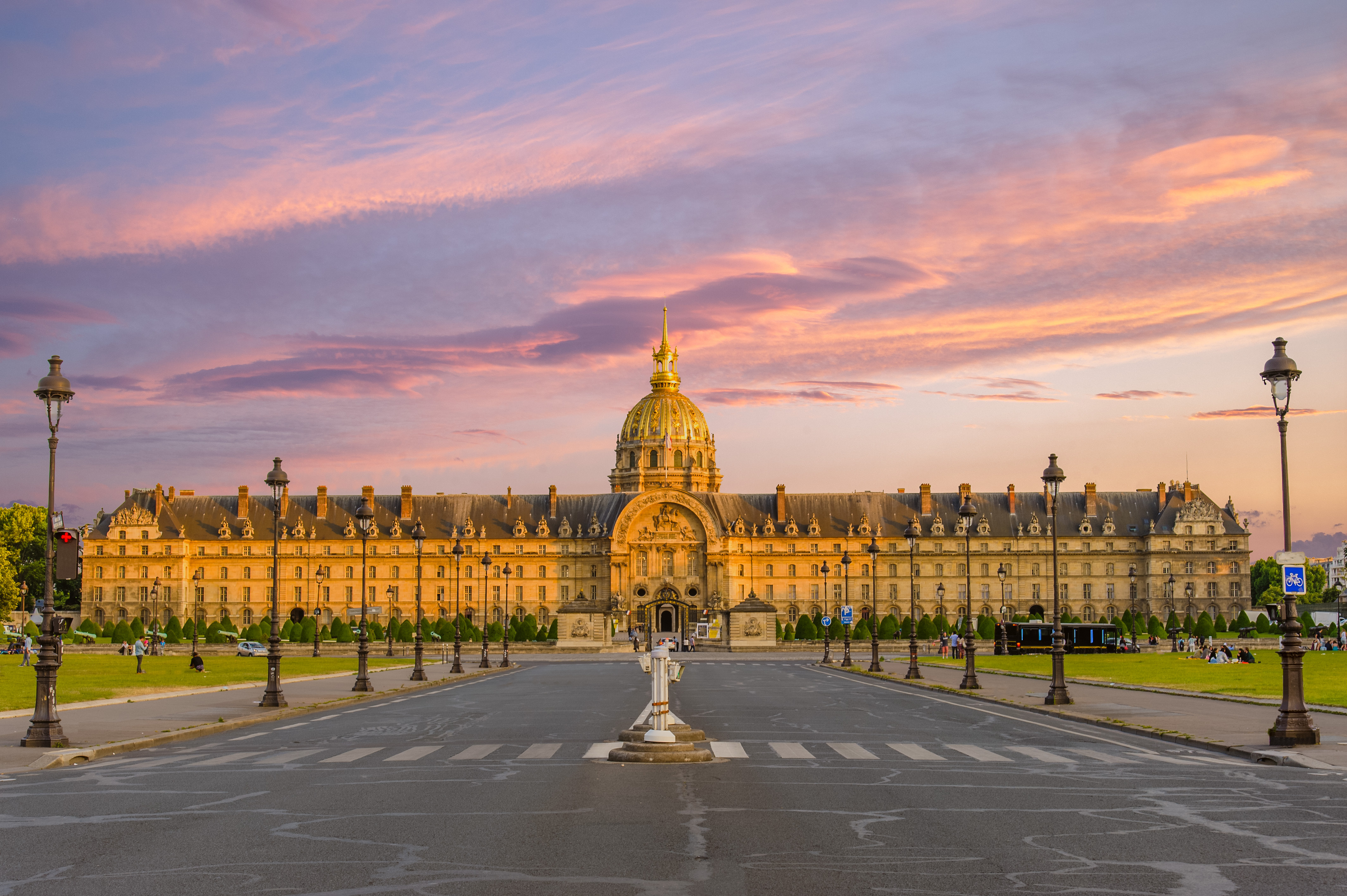 Bâtiment historique avec un grand dôme doré au centre sous un ciel au coucher du soleil aux teintes roses et violettes, entouré d'une pelouse verte et de lampadaires en ligne sur une large avenue vide.