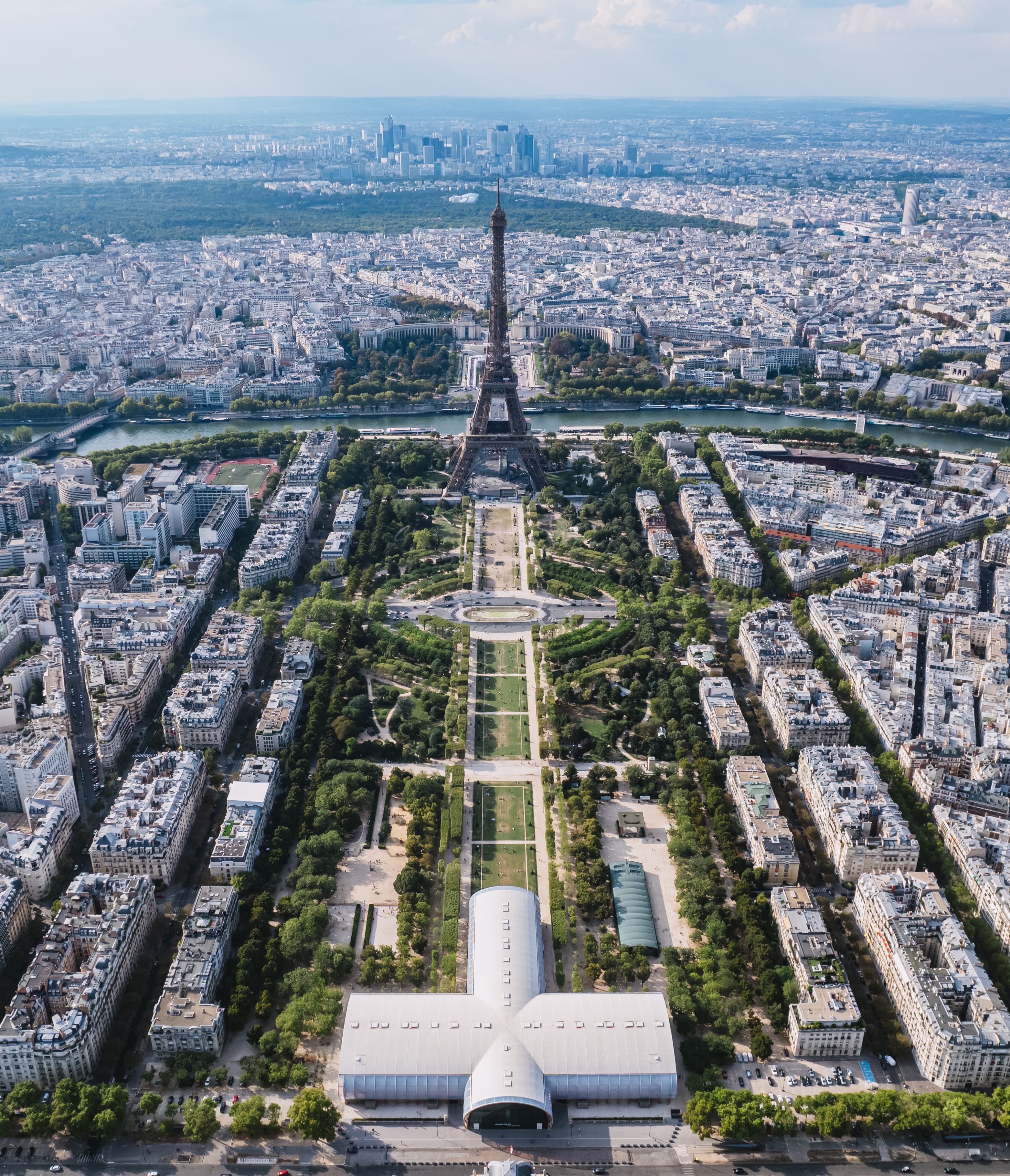 Vue aérienne de la tour Eiffel à Paris, entourée de jardins et de bâtiments urbains, avec la Seine au milieu et des gratte-ciel à l'horizon.