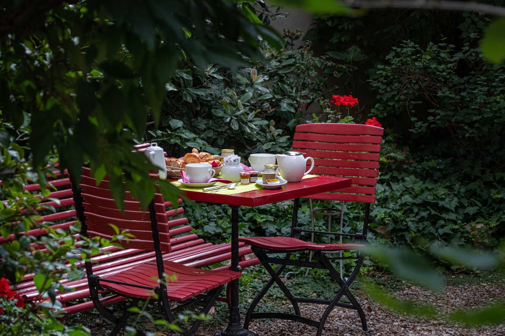 Table rouge avec petit-déjeuner composé de thé, confitures, viennoiseries et tasses, entourée de chaises rouges dans un jardin verdoyant avec feuillage dense et fleurs rouges.