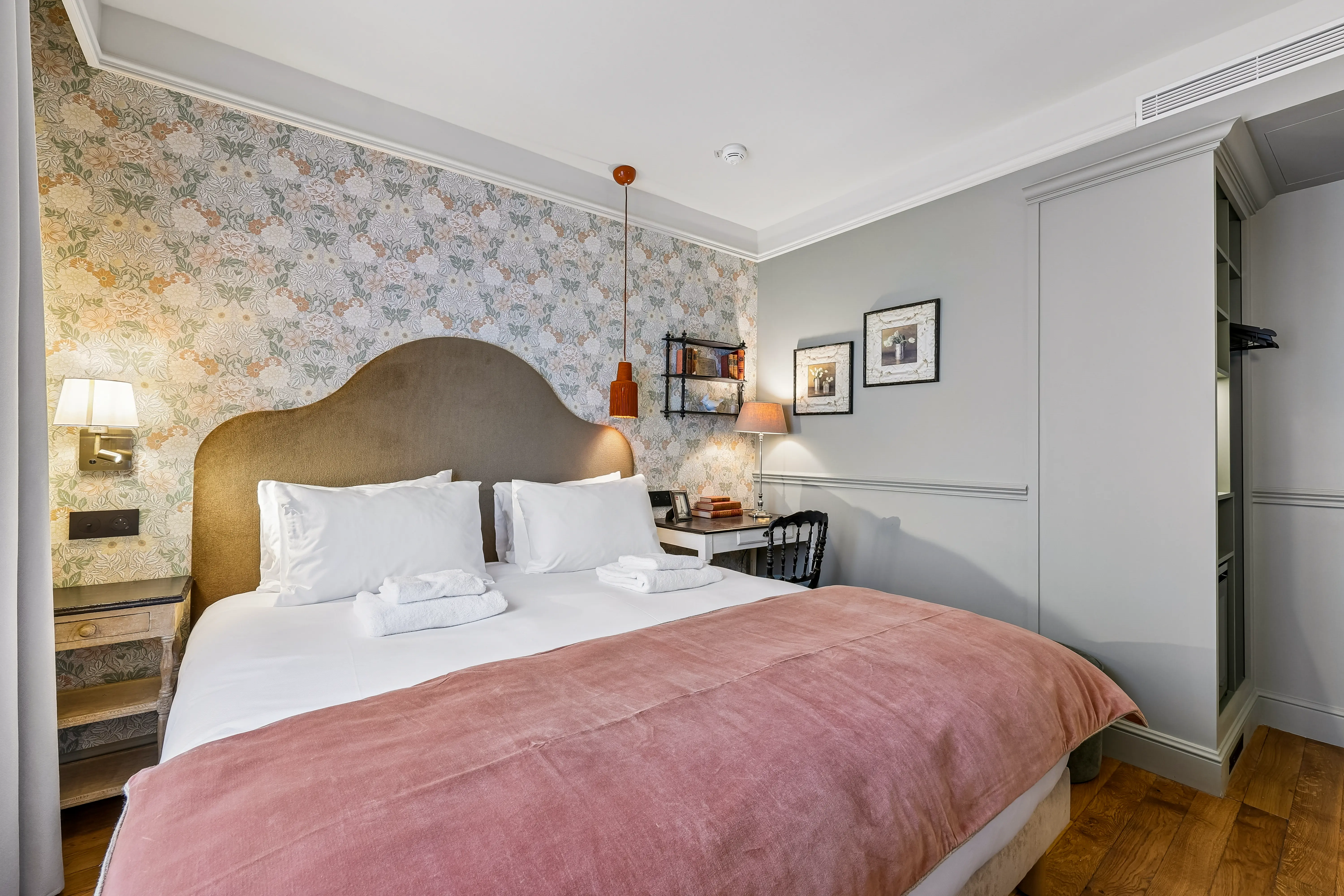 Chambre avec lit double avec tête de lit beige, couverture rose, deux oreillers blancs et serviettes pliées. Mur décoré avec papier peint floral d'un côté et peinture grise de l'autre. Table de nuit avec lampe murale à gauche, bureau blanc avec lampe de bureau, livres et chaise noire à droite. Deux cadres photos accrochés au mur gris. Sol en parquet.