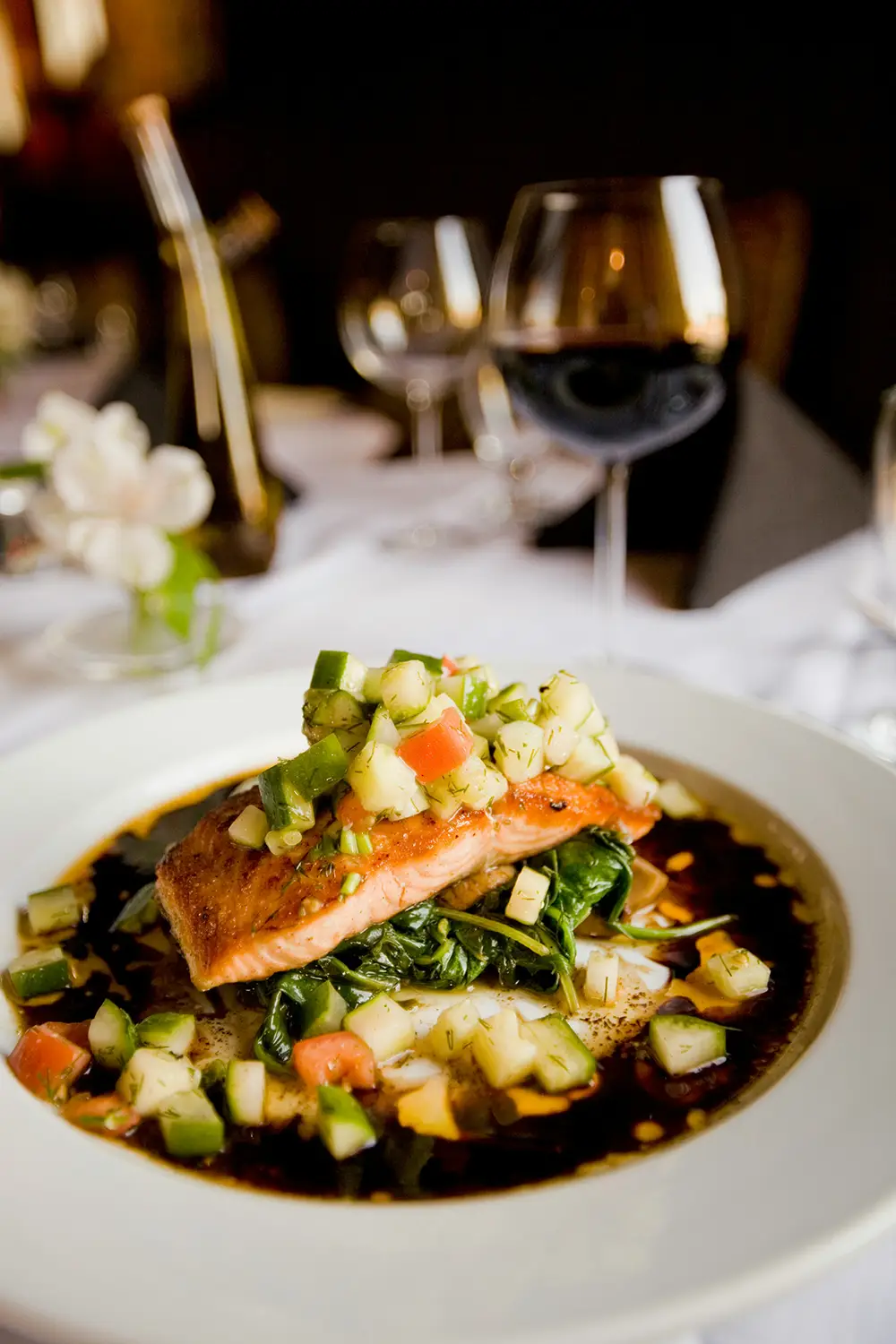 Plat raffiné avec filet de saumon grillé sur un lit d'épinards, décoré de dés de légumes frais et servi avec une sauce foncée, accompagné d'un verre de vin rouge sur une table élégamment dressée.
