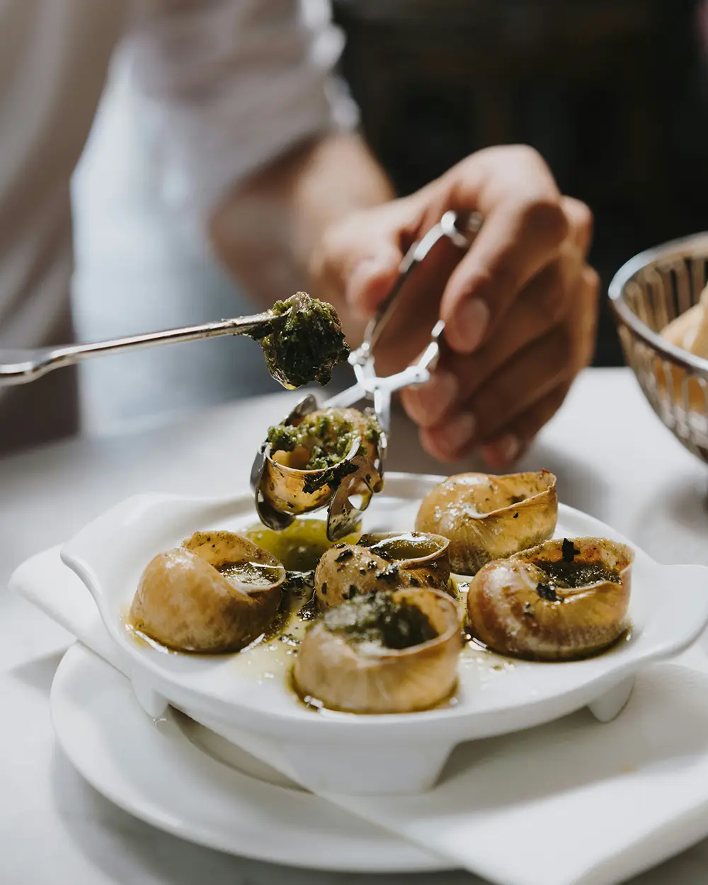Main tenant une pince spéciale pour escargots, soulevant un escargot avec du beurre à l'ail et au persil à côté d'autres escargots servis dans une assiette blanche creuse.