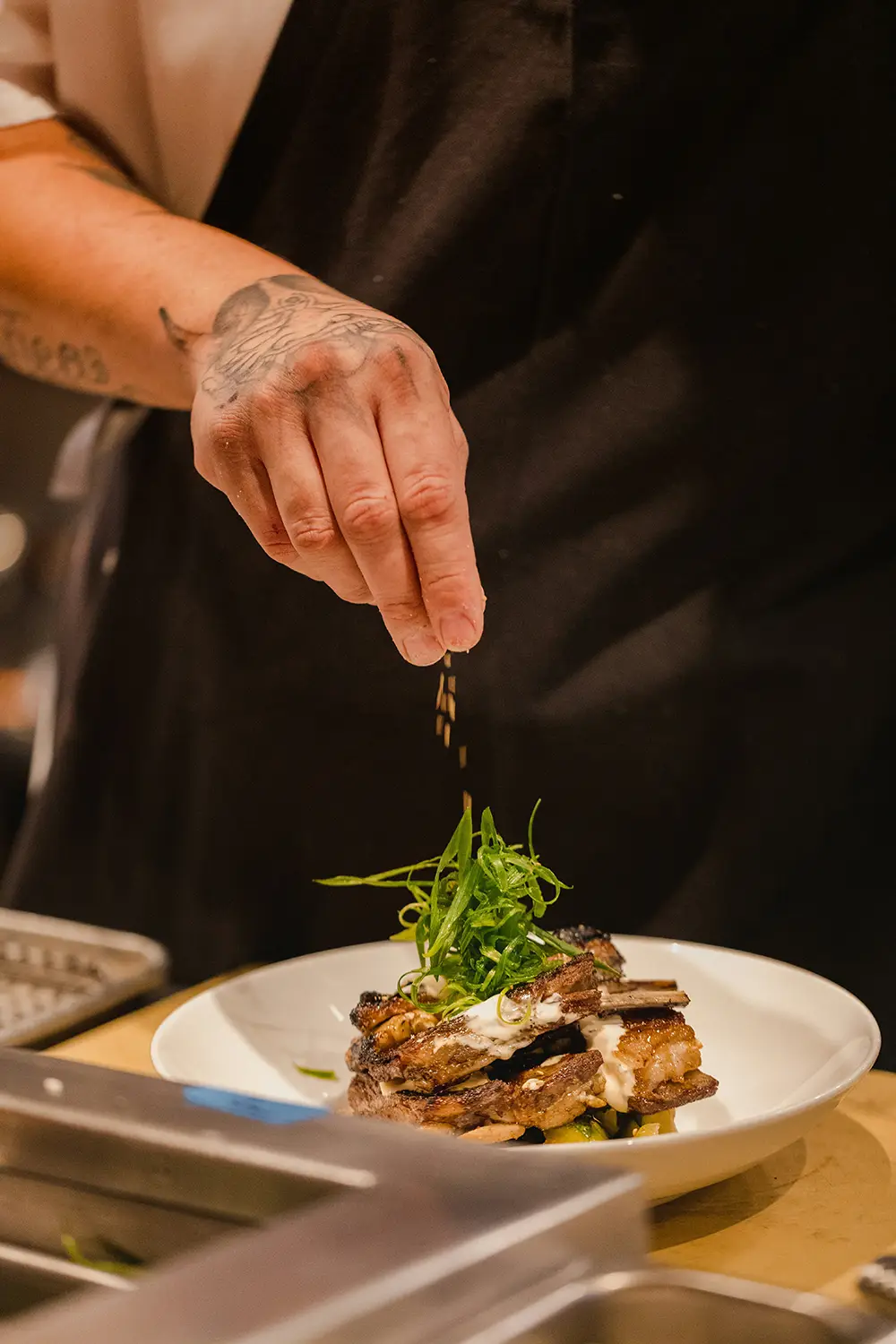Main cuisinier avec un tatouage sur la main qui saupoudre des épices sur des côtes grillées garnies de verdure dans une assiette blanche.