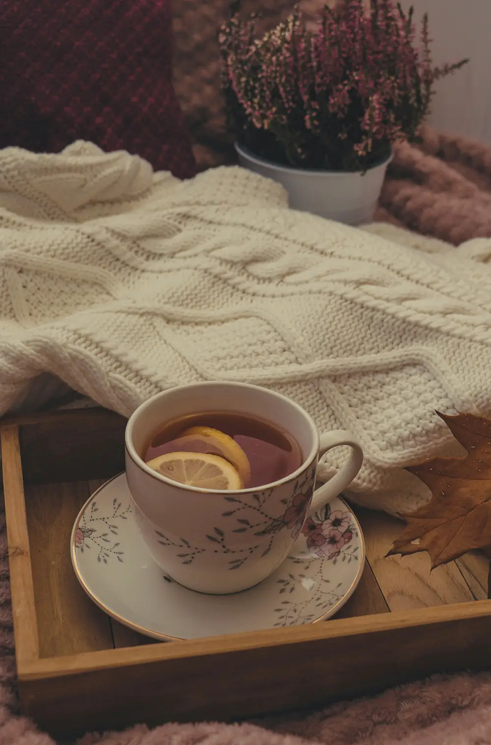Tasse de thé avec des rondelles de citron sur une soucoupe décorée, posée dans un plateau en bois sur une couverture en laine crème, avec une plante en pot à fleurs violettes et une feuille d'automne marron à côté.