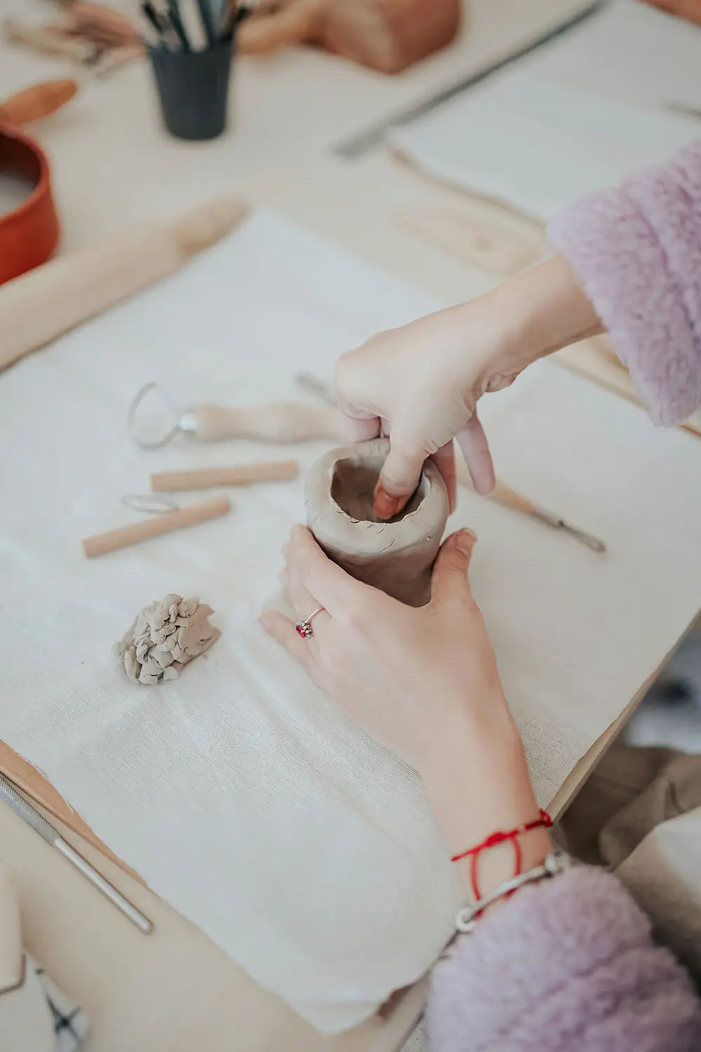 Une personne façonne une poterie en argile avec ses mains sur une table couverte d'un tissu blanc, entourée d'outils de poterie en bois.