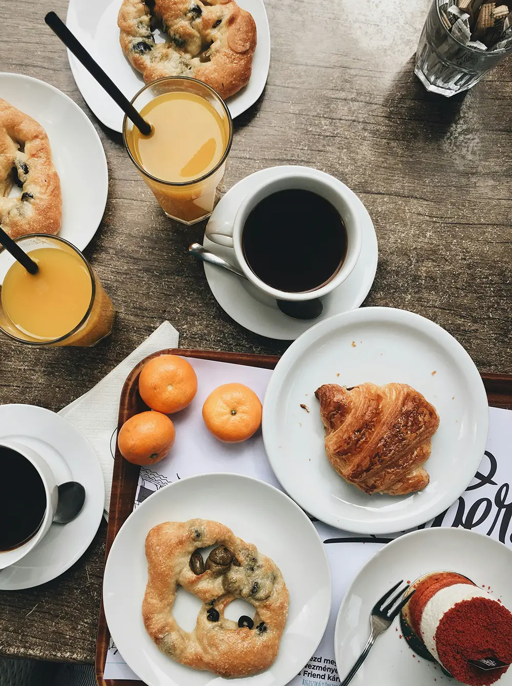 Une table en bois avec deux tasses de café noir, deux verres de jus d'orange avec pailles, un croissant, des petits pains aux olives sur des assiettes blanches, trois mandarines et un dessert pâtissier à base de crème et de poudre rouge.