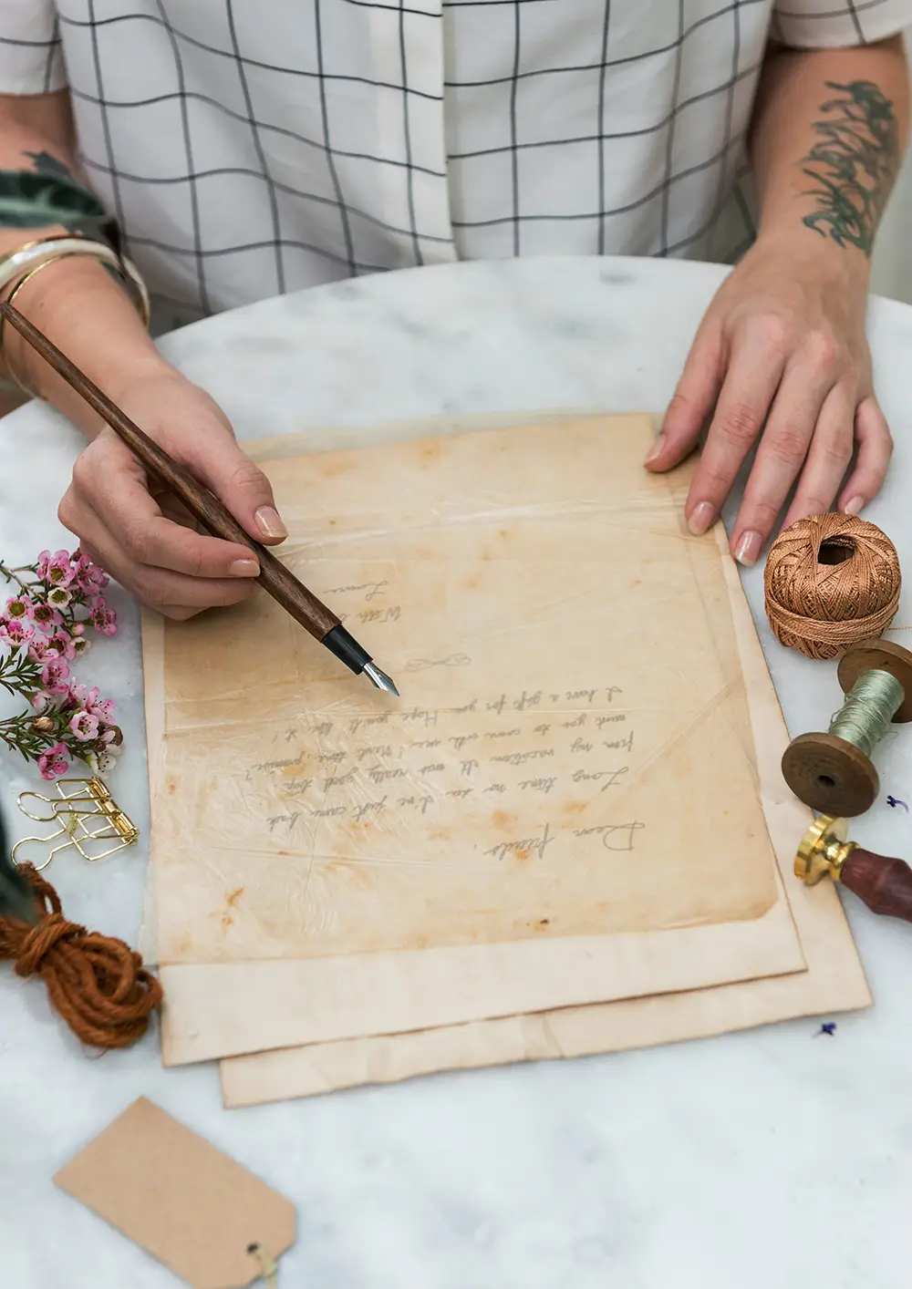 Une personne en chemise blanche à carreaux tient un stylo plume en bois au-dessus d'une lettre ancienne jaunie posée sur une table en marbre, entourée d'objets comme une bobine de fil, fleurs roses, pince à papier dorée et un cachet de cire.