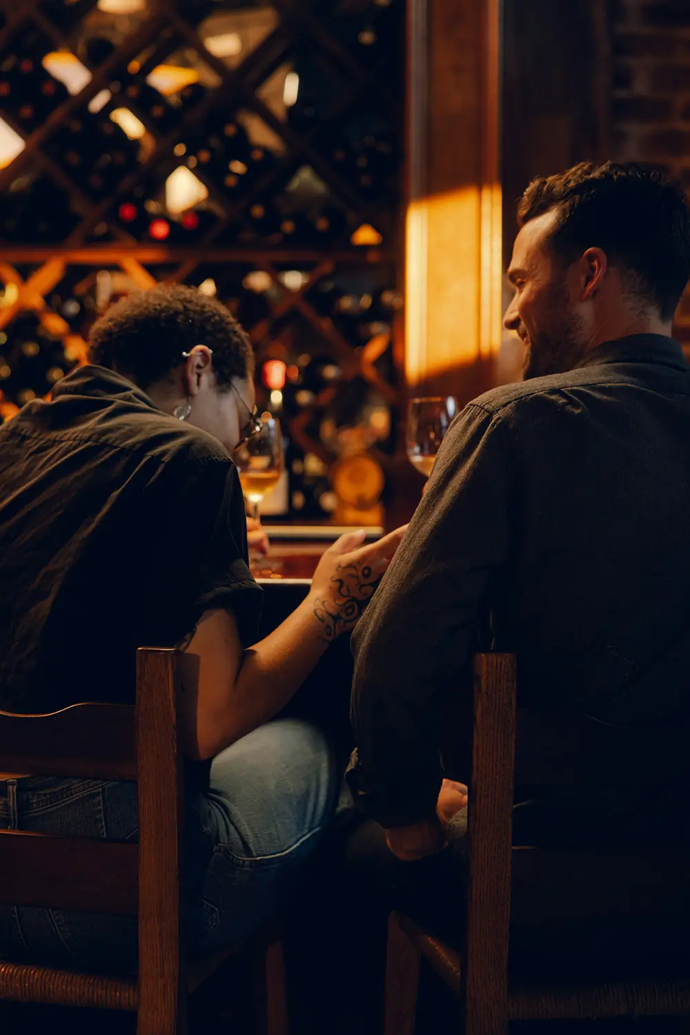 Deux personnes assises dos à dos au bar d'une cave à vin, partageant un moment convivial avec des verres de vin devant elles.