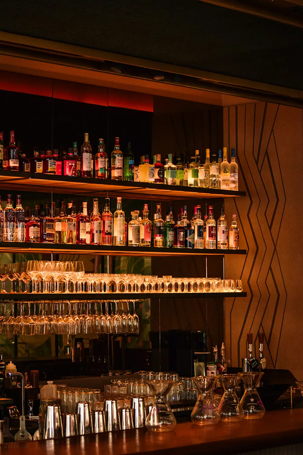 Bar avec étagères en bois alignant diverses bouteilles d'alcool et plusieurs rangées de verres à pied et verres à cocktail suspendus, éclairage chaleureux.