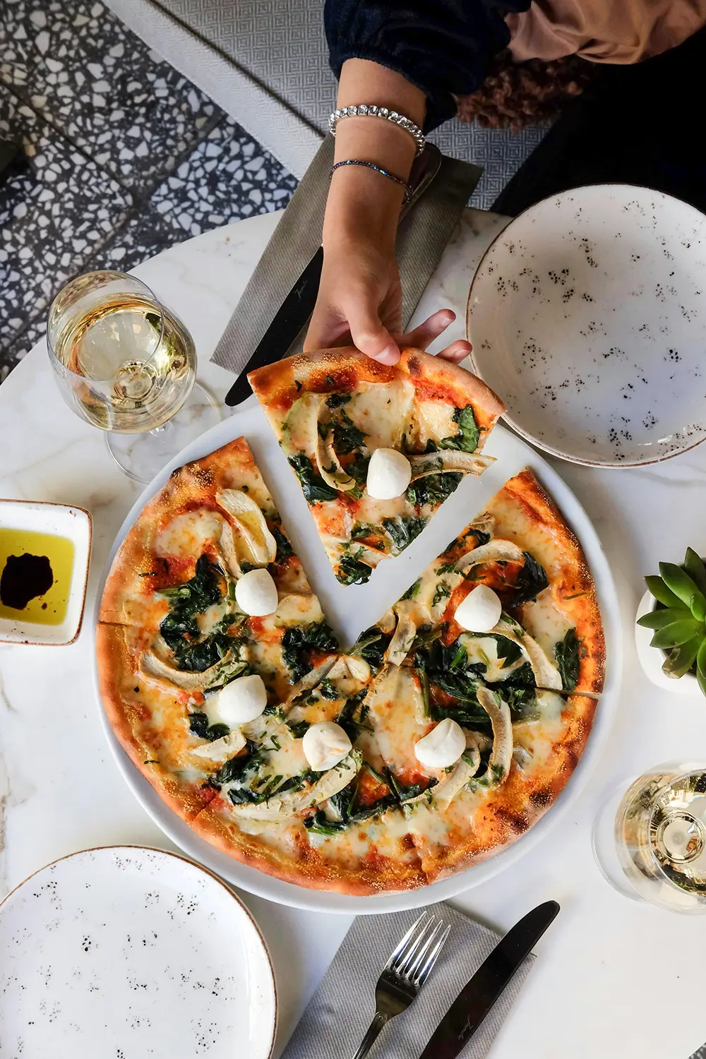Une main avec deux bracelets tient une tranche de pizza sur une table avec une pizza garnie de fromage, épinards, champignons et boules de mozzarella, deux verres de vin blanc, un petit plat d'huile et de vinaigre, des assiettes blanches à motifs et des couverts sur des serviettes grises.