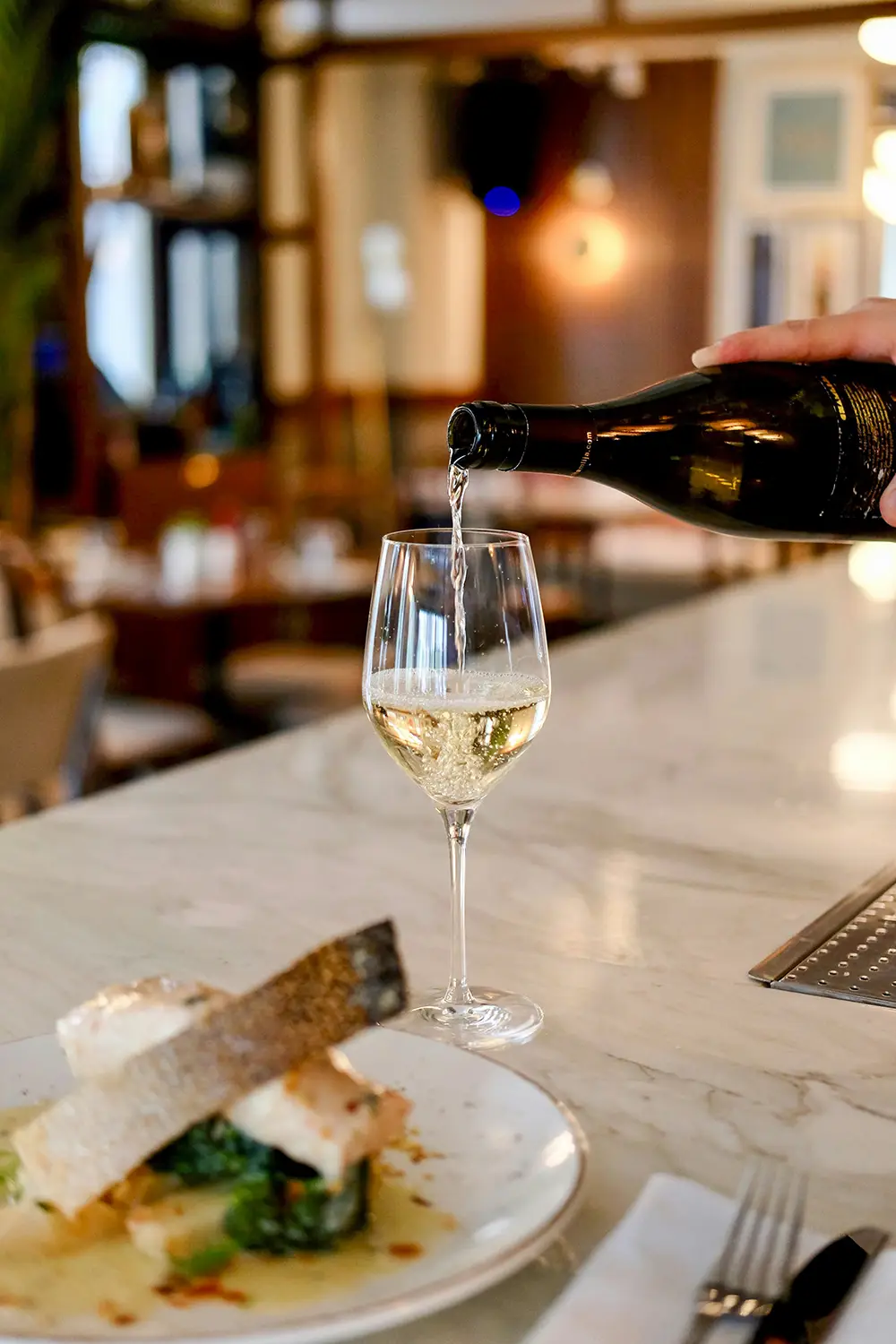 Un verre de vin blanc est rempli à partir d'une bouteille noire tenue par une main sur un comptoir en marbre, avec une assiette de poisson et légumes en premier plan dans un restaurant.