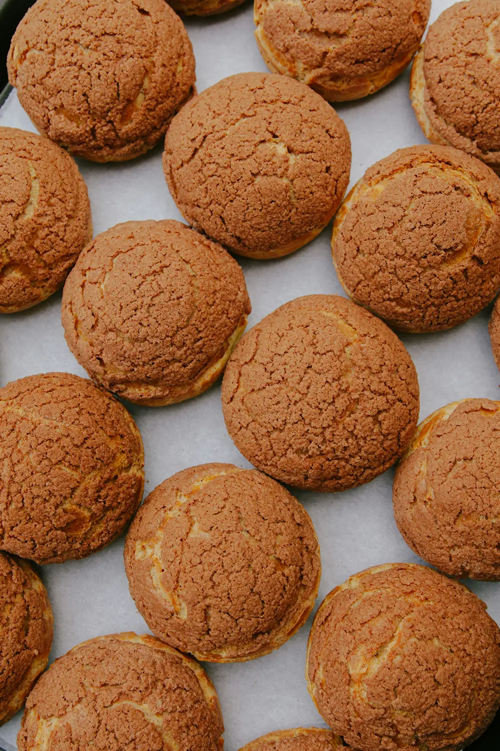 Des choux à la crème cuits, avec une croûte craquelée dorée, disposés en rangées sur du papier sulfurisé.