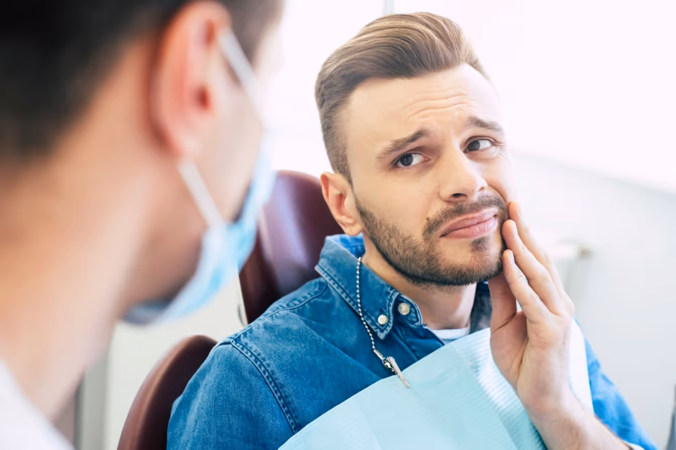 Man holding his cheek and expressing tooth pain while speaking with a dentist