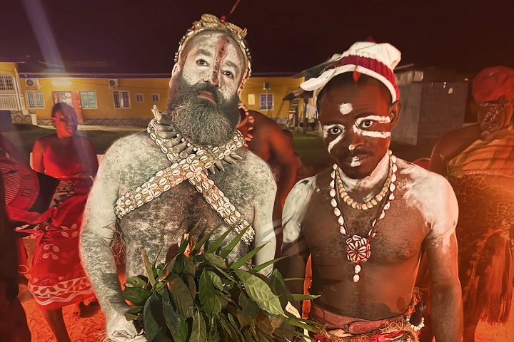 Two people with body paint and traditional shell necklaces pose at a nighttime cultural celebration, with one holding green leaves and other participants in the background